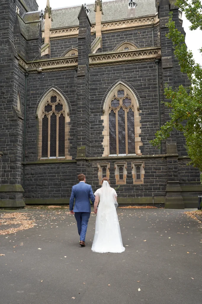 emily declan wedding st patricks cathedral courtyard recessional This image captures a bride and groom walking away from the camera in a paved courtyard, with St Patrick's Cathedral in Melbourne as the dramatic historic backdrop. They walk hand-in-hand, the bride in a white lace wedding dress with a long train and veil, and the groom in a light blue suit. The scene conveys timeless elegance, solemnity, and quiet joy, with the dark Gothic Revival bluestone cathedral contrasting beautifully against the couple. Fallen leaves and surrounding foliage add a natural, seasonal touch to this serene and intimate wedding moment.