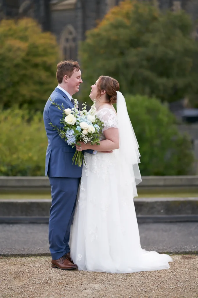 emily declan wedding st patricks cathedral garden portrait This image captures a joyful, intimate portrait of a bride and groom standing on a gravel path in a garden area at St Patrick's Cathedral in Melbourne. The bride wears a white V-neck A-line lace gown with a flowing veil and holds a white and blue bouquet, while the groom is in a light blue suit and brown shoes. They face each other, smiling and laughing, with the cathedral softly blurred in the background, creating a romantic, elegant, and serene wedding moment.