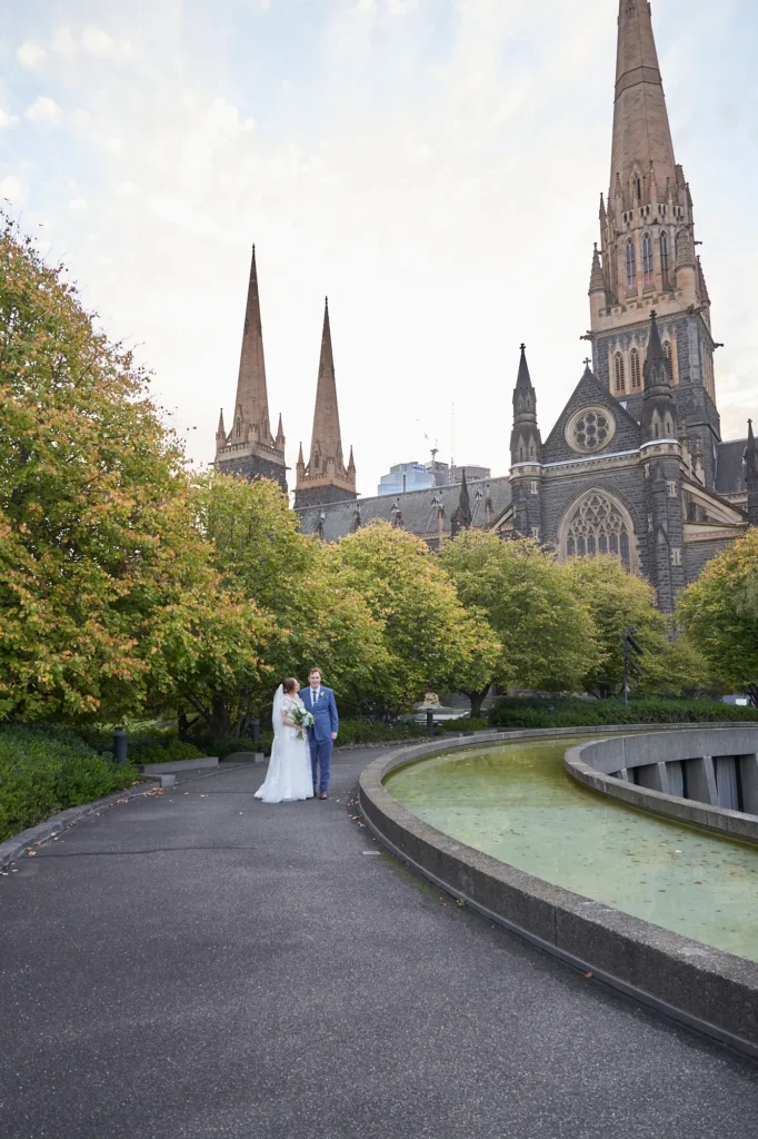 emily declan wedding st patricks cathedral park walk This image captures a bride and groom walking together along a paved path in a park area with St Patrick's Cathedral in Melbourne as the dramatic backdrop. The bride wears a white lace V-neck A-line gown with a train, and the groom is in a light blue suit. The Gothic Revival cathedral with soaring spires and intricate stonework contrasts with the natural greenery and a curved water feature in the foreground, creating a grand, serene, and historic atmosphere for this wedding portrait.
