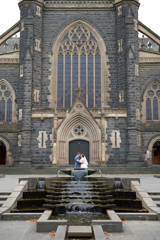 emily declan wedding st patricks cathedral water feature portrait This image captures a bride and groom posing near a large stone water feature in front of St Patrick's Cathedral in Melbourne. The couple stands side-by-side, looking at each other, with the bride in a white lace V-neck A-line gown and the groom in a light blue suit. The dark bluestone Gothic Revival cathedral, intricate stonework, tall pointed-arch windows, and flowing water in the foreground create a grand, serene, and historic backdrop. Fallen autumn leaves on the paved area enhance the timeless and intimate atmosphere of this wedding portrait.