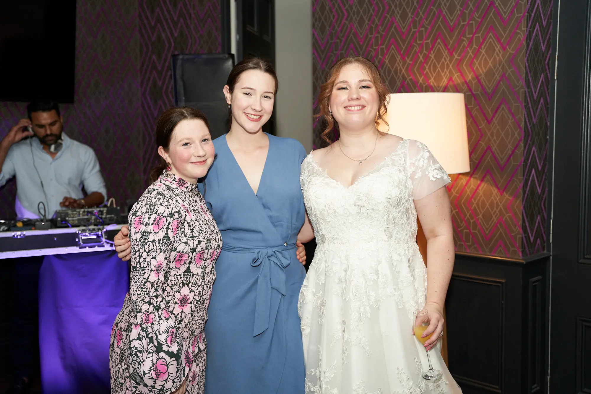 emily declan wedding the vincent albert park bride and guests group portrait The bride shares a joyful group portrait with two female guests at The Vincent, Albert Park. The bride wears a white V-neck lace dress with cap sleeves, holding a glass of champagne, while the guests wear a dusty blue wrap dress and a floral patterned dress with pink earrings. All three smile warmly at the camera, standing closely together. The background features purple geometric wallpaper, a floor lamp, and a DJ booth with ambient and purple uplighting, creating a vibrant, celebratory, and elegant wedding reception atmosphere.
