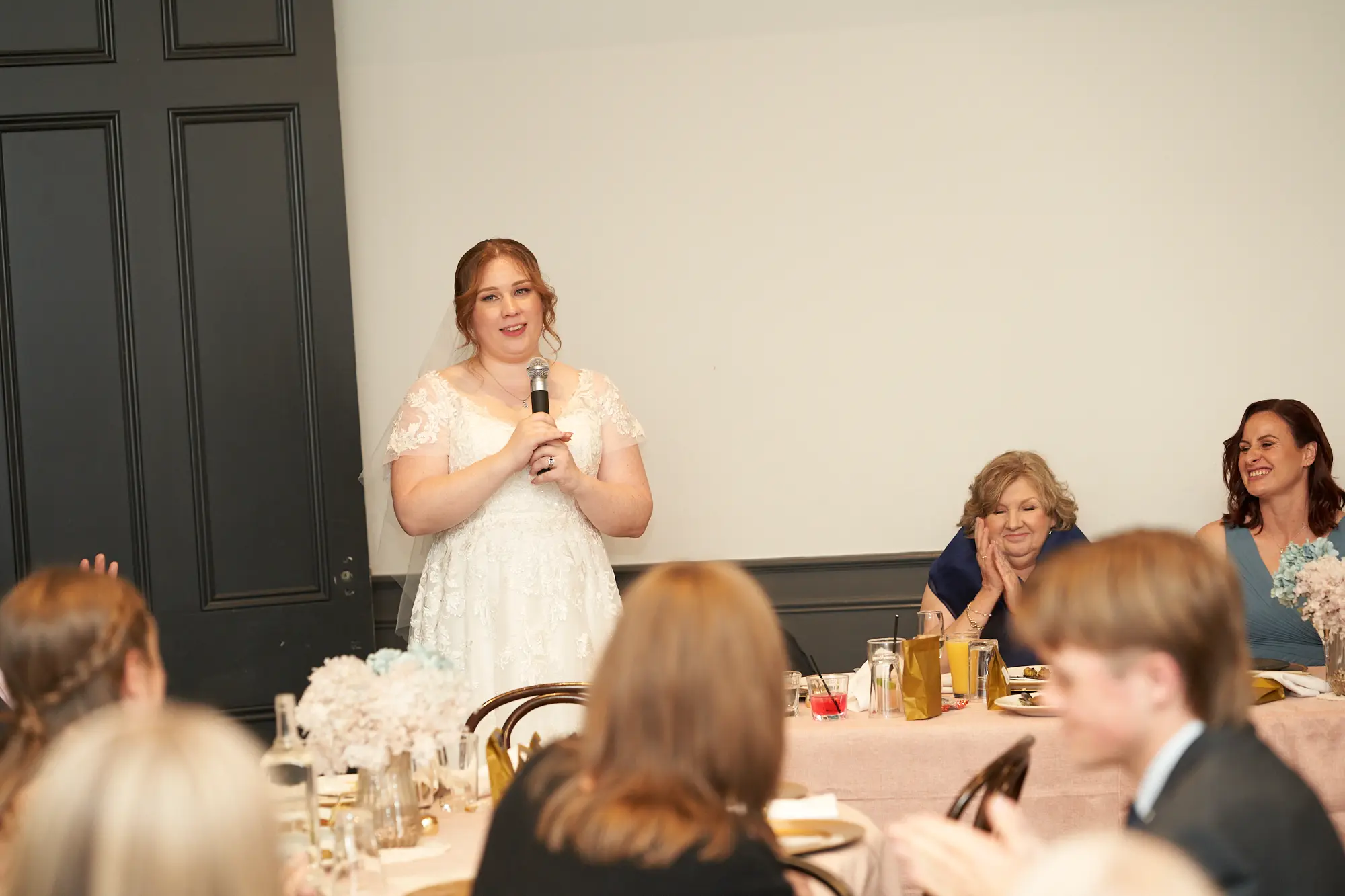 emily declan wedding the vincent albert park bride giving speech The bride gives a heartfelt speech at her wedding reception at The Vincent, Albert Park, holding a microphone and smiling warmly. Seated guests, including women in navy blue dresses, listen attentively in the background. Tables in the foreground are covered with pale pink tablecloths and decorated with gold-accented plates, glassware, and white and pale blue floral arrangements. The indoor space features a plain white wall, tall black wooden doors, high ceilings, and elegant trim. The scene captures a joyful, intimate, and engaging moment, highlighting the warmth and elegance of the celebration.