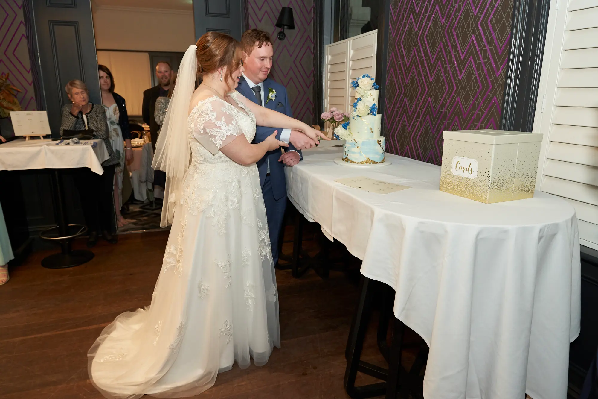 emily declan wedding the vincent albert park cake cutting moment The bride and groom share a joyful moment cutting their three-tiered wedding cake at The Vincent, Albert Park. They stand side-by-side, the groom’s arm around the bride as they both hold the cake knife. Guests watch from the background, smiling and engaged. The venue features purple geometric wallpaper, white louvered shutters, and dark wood accents. The white-covered table holds the cake and a gift box labeled "Cards." The image captures the elegance, happiness, and intimate celebration of this classic wedding tradition in a stylish indoor setting.