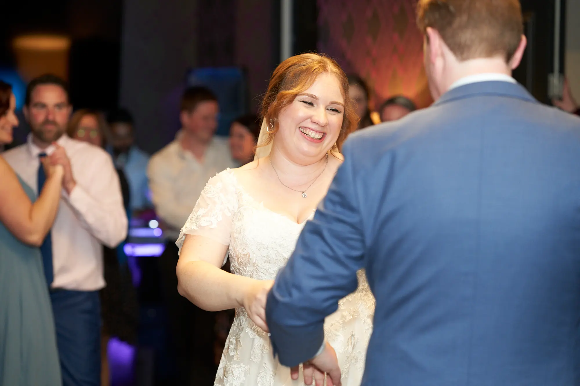 emily declan wedding the vincent albert park first dance candid The bride and groom share a joyful, intimate first dance at The Vincent, Albert Park. They are in a close embrace, smiling at each other as nearby guests watch or dance along. The bride wears a white V-neck lace dress with cap sleeves, and the groom wears a light blue suit with a patterned tie. Warm, ambient lighting highlights the couple against the dimly lit room with purple geometric wallpaper and subtle bokeh from accent lights, creating a romantic, celebratory, and elegant atmosphere at this special wedding reception moment.