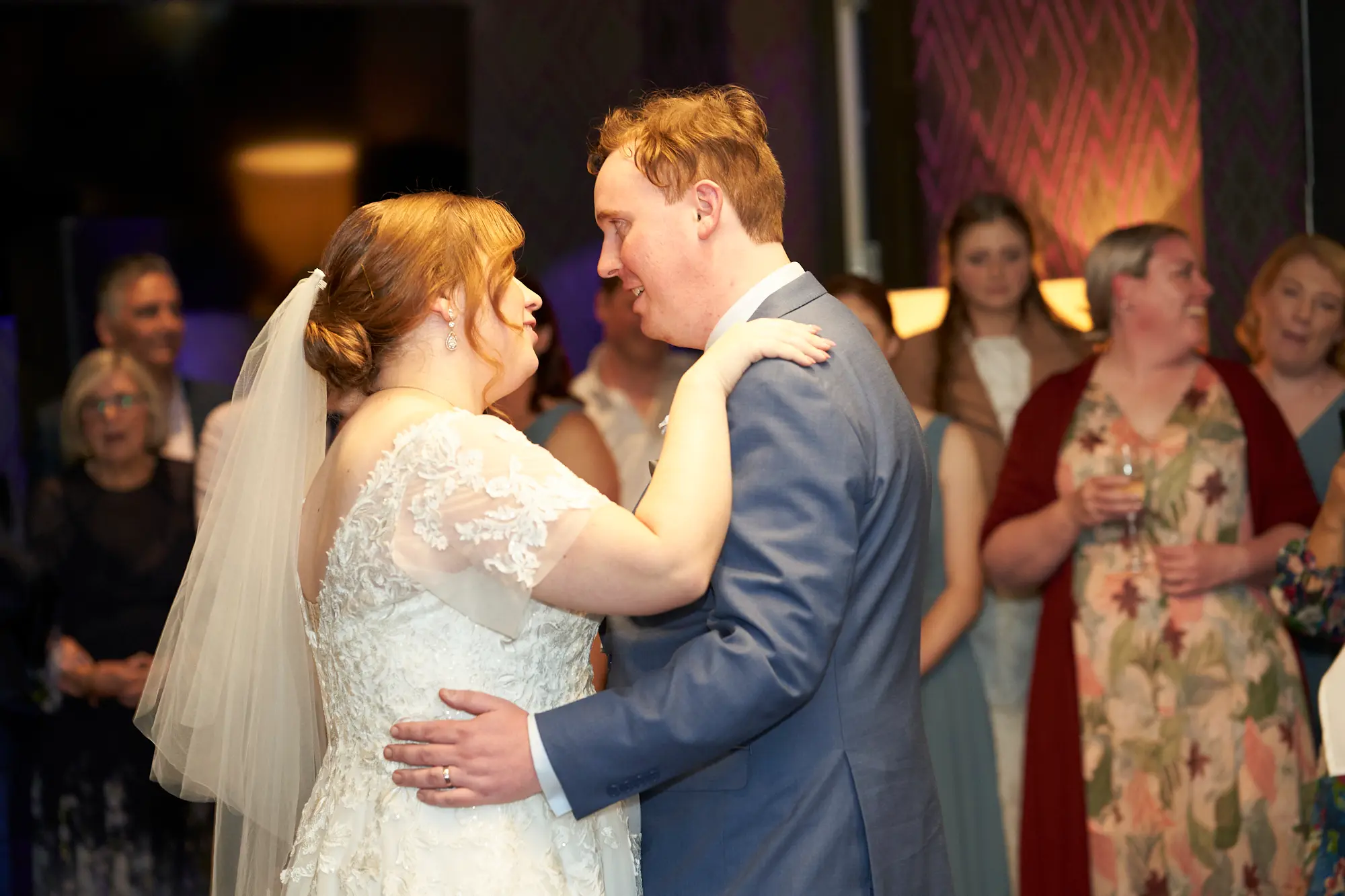 emily declan wedding the vincent albert park first dance The bride and groom share a joyful, intimate first dance at The Vincent, Albert Park. They are in a close embrace, smiling at each other as guests look on in the softly blurred background. The bride wears a white V-neck lace dress with cap sleeves, and the groom wears a light blue suit with a patterned tie. Warm ambient lighting and a dimly lit room with purple geometric wallpaper and dark wood accents create a romantic and celebratory atmosphere, capturing the love and happiness of this special wedding reception moment.