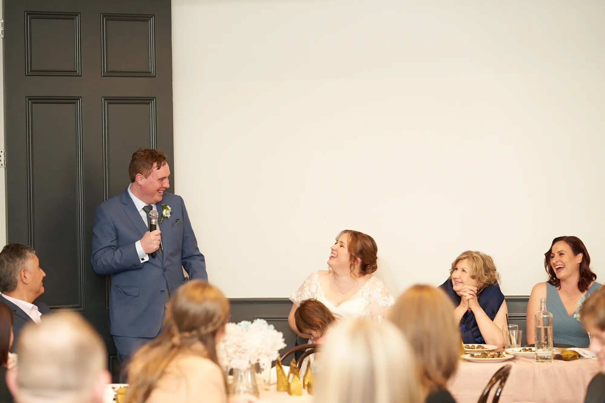 emily declan wedding the vincent albert park groom speech candid moment The groom delivers a lively speech at his wedding reception at The Vincent, Albert Park, holding a microphone and smiling widely. The bride sits beside him, laughing with her hand resting on his arm. Guests at the head table, including a man in a navy suit and two women in blue dresses, are engaged and smiling. The indoor space features pale pink tablecloths, white floral arrangements, a tall dark gray door, and a purple geometric wallpaper. The image captures a joyful, intimate, and celebratory moment, highlighting the warmth and elegance of the reception.