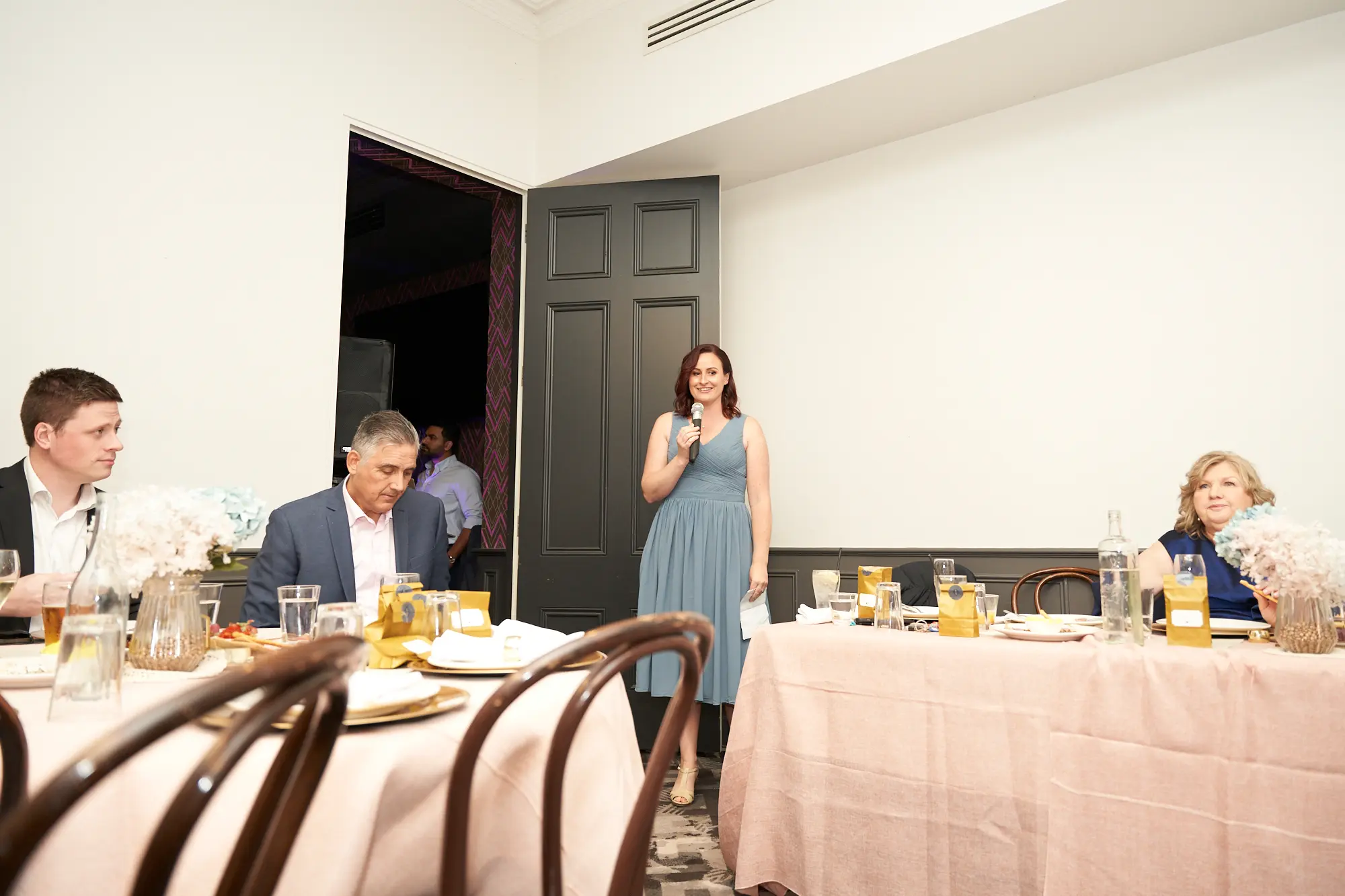emily declan wedding the vincent albert park reception bridesmaid speech A bridesmaid gives a heartfelt speech during a wedding reception at The Vincent in Albert Park. She stands on the right, holding a microphone and gesturing as she speaks, wearing a dusty blue V-neck knee-length dress. Guests seated at the head table listen attentively, with wine glasses, gold-accented plates, and soft floral arrangements decorating the pale pink tablecloth. A white wall and dark paneled doorway with geometric purple wallpaper form the backdrop, creating an intimate and celebratory atmosphere.