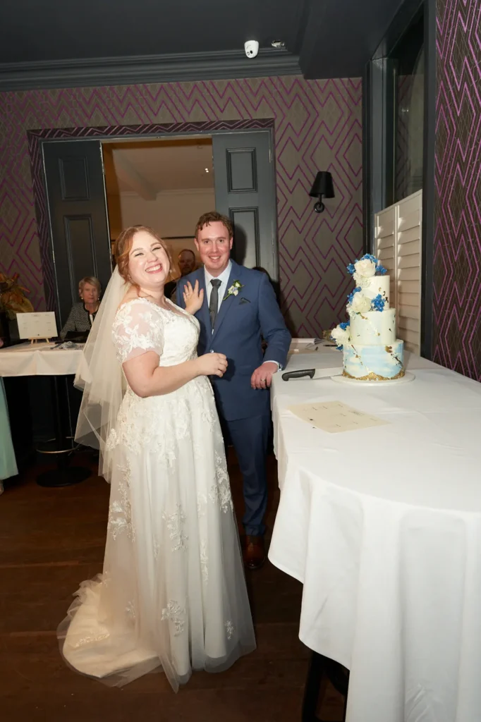 emily declan wedding the vincent albert park reception cake moment A joyful candid portrait of the bride and groom during their reception at The Vincent in Albert Park. The couple stand beside a long white-tableclothed table with their three-tiered wedding cake and signing documents. The bride smiles brightly toward the camera in her V-neck lace gown and veil, while the groom, in a light blue suit, smiles beside her. The background features geometric purple wallpaper, warm wood flooring, and soft indoor lighting, creating an elegant and celebratory atmosphere.