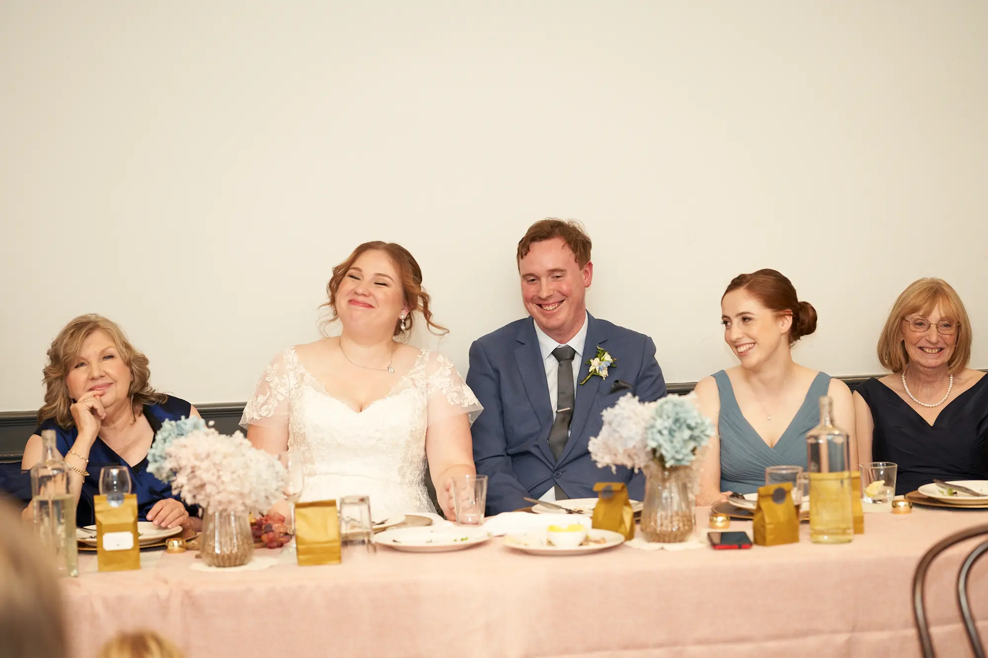 emily declan wedding the vincent albert park reception head table laughter moment A joyful candid moment at the wedding reception head table at The Vincent in Albert Park, showing the bride laughing openly while the groom smiles warmly beside her. An older woman sits to the bride’s left, smiling with her hand near her face, while two women on the groom’s right look on with gentle smiles. The long table is decorated with a pale pink cloth, gold-accented plates, glassware, and floral arrangements in white, pink, and pale blue. A simple white wall and warm indoor lighting create an elegant, intimate atmosphere.