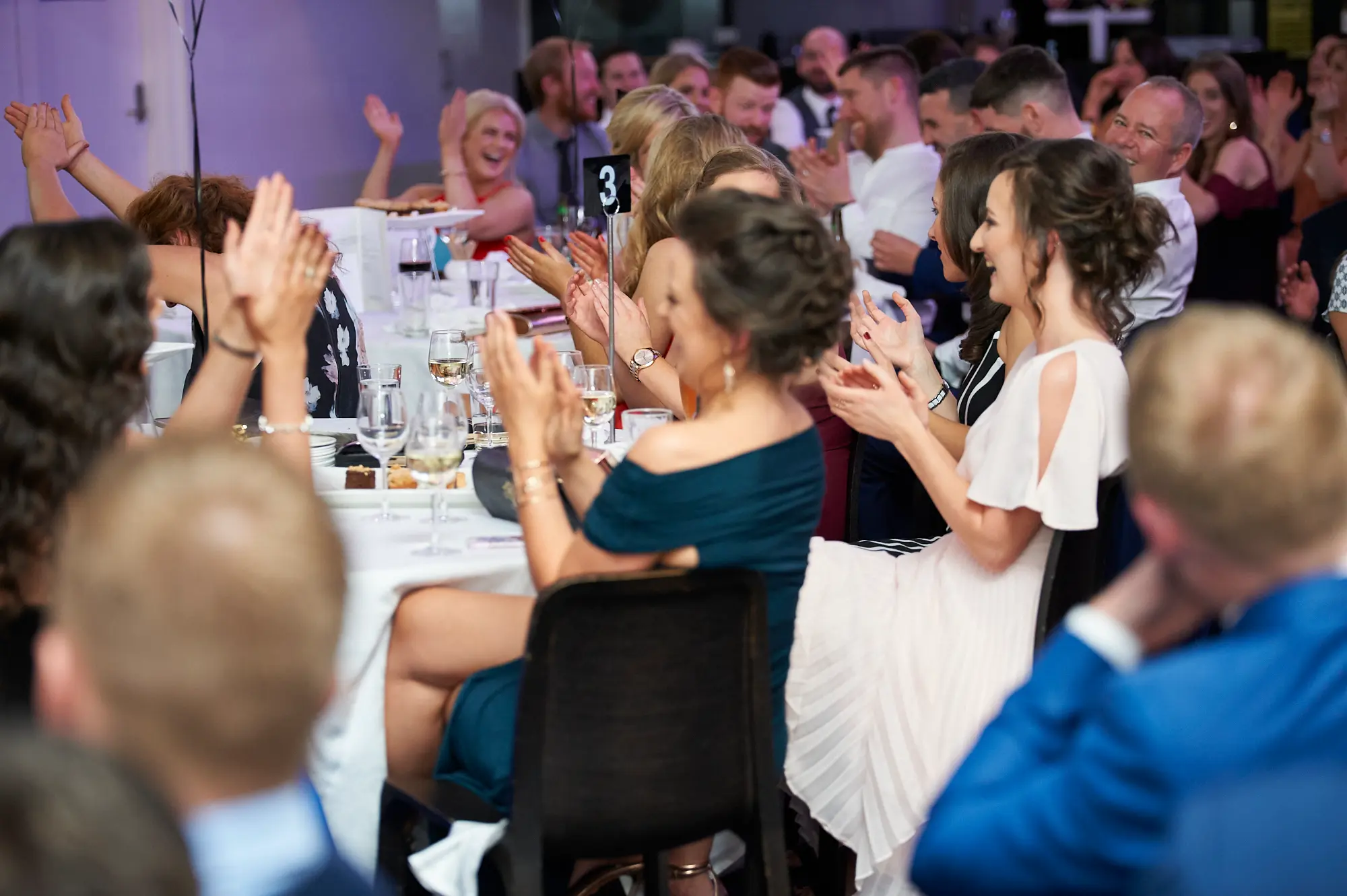 padraig pearses gac awards night mcg applause celebration tables Guests at the Pearses Awards Night at the MCG are clapping and celebrating while seated at round dinner tables. A woman in a dark teal off-the-shoulder dress and another in a white and pink dress applaud enthusiastically, smiling warmly. In the background, guests in red and blonde dresses laugh and clap, all looking toward a stage or presentation. White tablecloths, wine glasses, plates, and a table number sign “3” set a formal dining scene. The warm ambient lighting and joyful expressions capture a lively, festive, and engaging moment of applause and recognition during the gala dinner.
