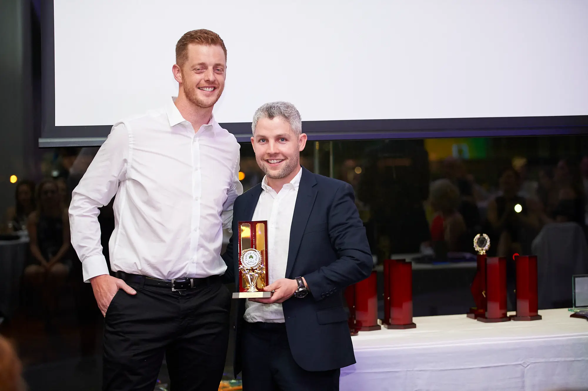 padraig pearses gac awards night mcg award posing Two men pose for a photograph at the Padraig Pearses GAC Awards Night at the MCG. The taller man in a white collared shirt and black trousers stands with one hand in his pocket, while the other man, in a dark suit jacket, proudly holds a golden trophy with a wooden base. Behind them, a large blank projection screen and a white-clothed table with multiple awards set the scene for a formal and celebratory moment. Warm ambient lighting and glimpses of seated guests in the background highlight the festive atmosphere of the gala awards dinner.