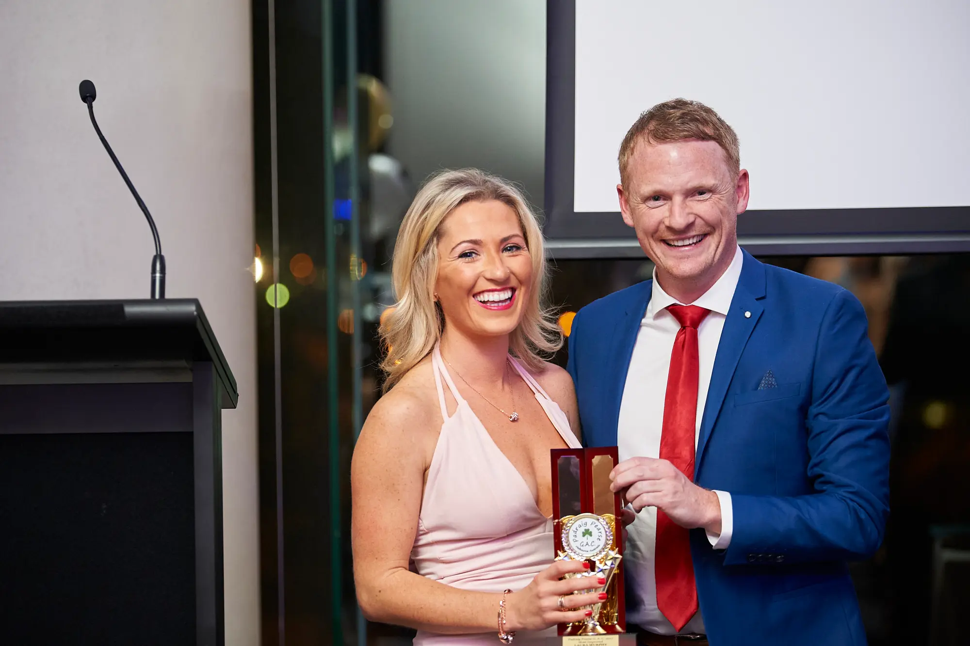padraig pearses gac awards night mcg award presentation photo A man in a blue suit proudly holds a golden award with a red emblem while standing beside a woman in a light pink dress at the Pearses Awards Night at the MCG. Both smile warmly at the camera, commemorating the moment of achievement. The modern venue features a black lectern, blank white projection screen, and glass partitions with ambient blue and yellow lighting, emphasizing the formal, celebratory, and professional atmosphere of this awards presentation.