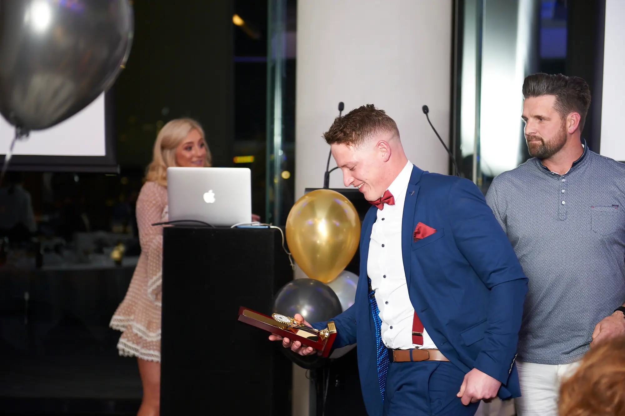 padraig pearses gac awards night mcg award presentation A man in a blue suit and red bow tie holds a wooden award at the Padraig Pearses GAC Awards Night at the MCG, smiling with pride. A colleague in a grey polo shirt stands supportively beside him, while a female host behind the lectern smiles brightly. Balloons and warm lighting highlight the celebratory, formal atmosphere of this awards presentation.