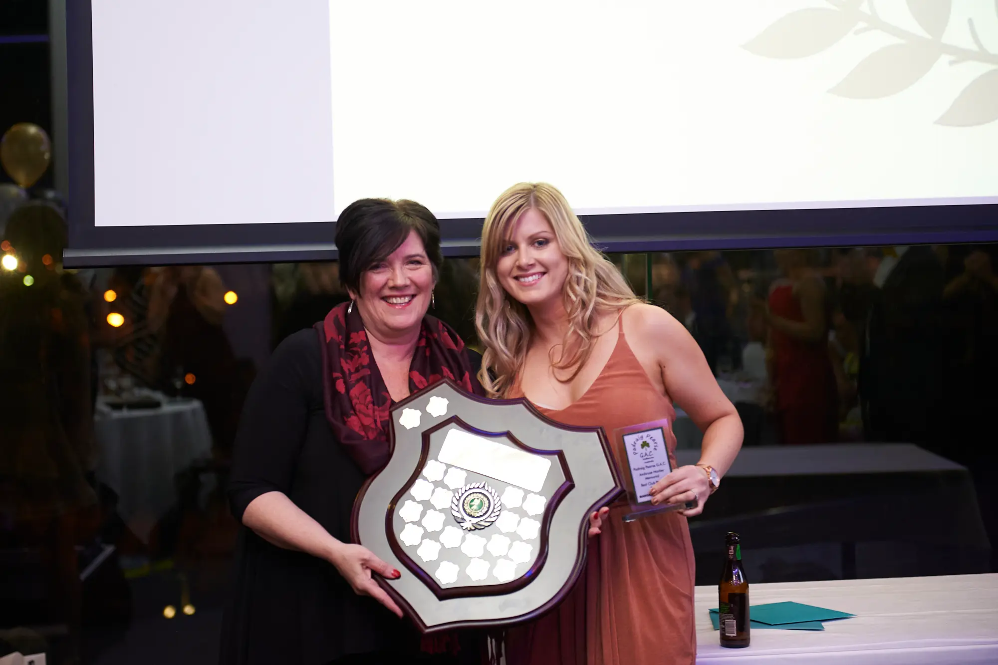 padraig pearses gac awards night mcg award recipients photo Two women stand side-by-side at the Pearses Awards Night at the MCG, smiling proudly while holding a large shield-style award and a smaller plaque. Both display broad, genuine smiles of accomplishment. The modern venue features white tablecloths, glass partitions, warm ambient lighting, and blurred guests in the background, creating a formal, celebratory, and engaging atmosphere that highlights the moment of recognition and professional achievement.