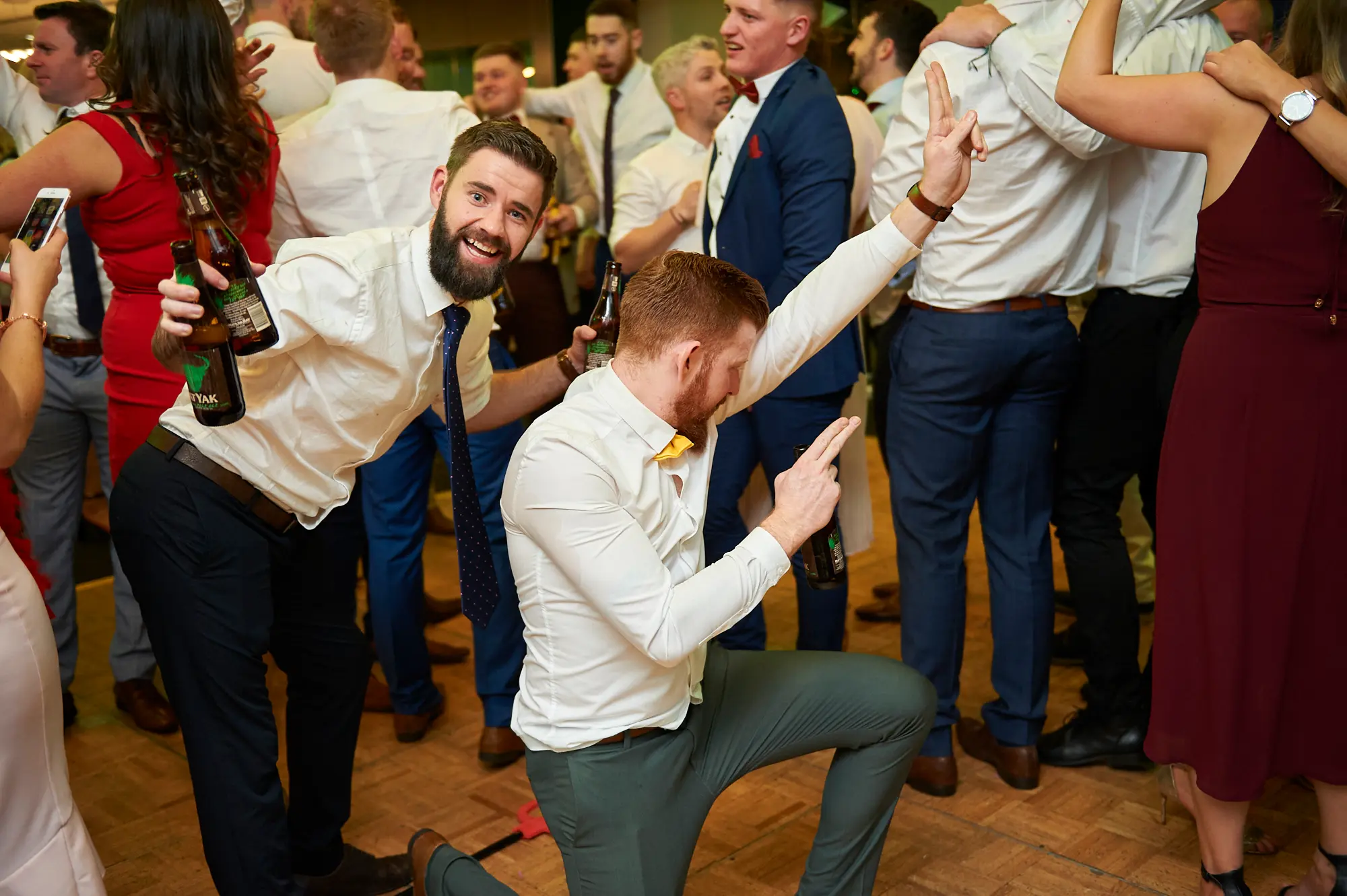padraig pearses gac awards night mcg beer celebration A man with red hair in a white shirt and yellow bow tie kneels on one knee, pointing and holding a beer bottle, while a bearded man in a white shirt and blue tie smiles broadly holding two beer bottles during the Pearses Awards Night at the MCG. Guests in formal attire cheer and raise their arms behind them on the wooden dance floor. The festive atmosphere is enhanced by warm ambient lighting, capturing a lively moment of camaraderie, joyful celebration, and spirited enjoyment at the gala event.