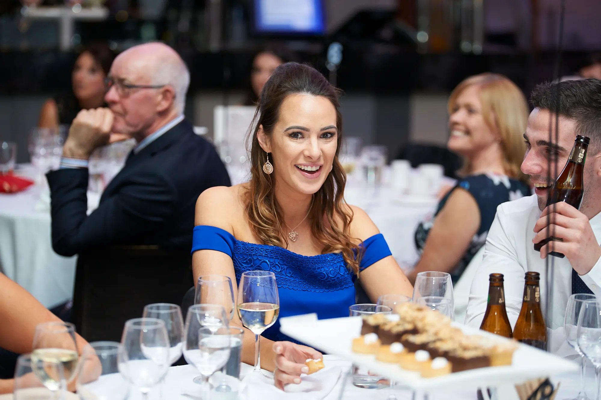 padraig pearses gac awards night mcg blue dress smile table Guests share a joyful moment at a dinner table during the Padraig Pearses GAC Awards Night & Gala Dinner at the MCG. A woman in a vibrant blue off-the-shoulder lace dress smiles directly at the camera, while a man beside her holds two brown glass beer bottles with an open-mouthed, lively expression. An older man and a woman in a floral dress are also visible, engaging with the table setting, which includes wine glasses, cutlery, and plates. Warm ambient lighting highlights the festive, social, and celebratory atmosphere of the gala event.