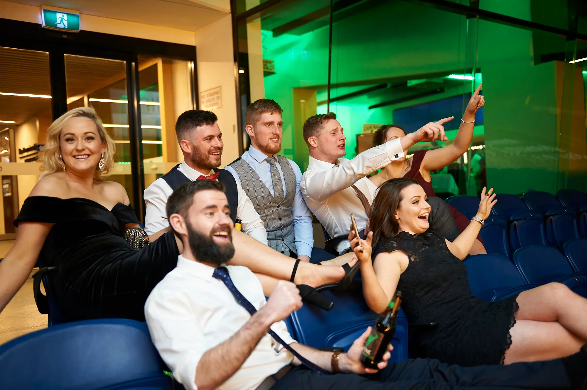 padraig pearses gac awards night mcg candid celebration guests cheering The image captures a lively candid moment at the Pearses Awards Night & Gala Dinner at the MCG, with guests in full celebration. A bearded man in a white shirt and tie gestures enthusiastically, mouth open in genuine excitement, holding a beer. A woman in a black dress raises her arm and phone, cheering exuberantly, while another man points animatedly off-camera. Nearby guests are engaged in the festive mood. The stadium seating and green ambient lighting provide a dynamic backdrop, emphasizing the electric, joyous atmosphere of a formal gala transformed into an energetic, spontaneous party.