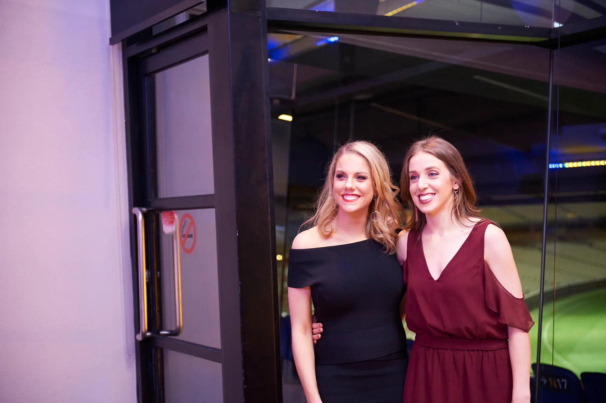 padraig pearses gac awards night mcg candid friends posing The image captures a candid moment of elegance and friendly posing at the Pearses Awards Night & Gala Dinner at the MCG. Two women stand side by side, smiling warmly at the camera, one in a black off-the-shoulder dress and the other in a burgundy cold-shoulder dress. Behind them, glass partitions and a large door reveal glimpses of blue stadium seating and vibrant blue ambient lighting, adding depth and sophistication to the modern venue. Their relaxed postures and engaging expressions convey a festive, social, and welcoming atmosphere, reflecting the celebratory nature of the evening.