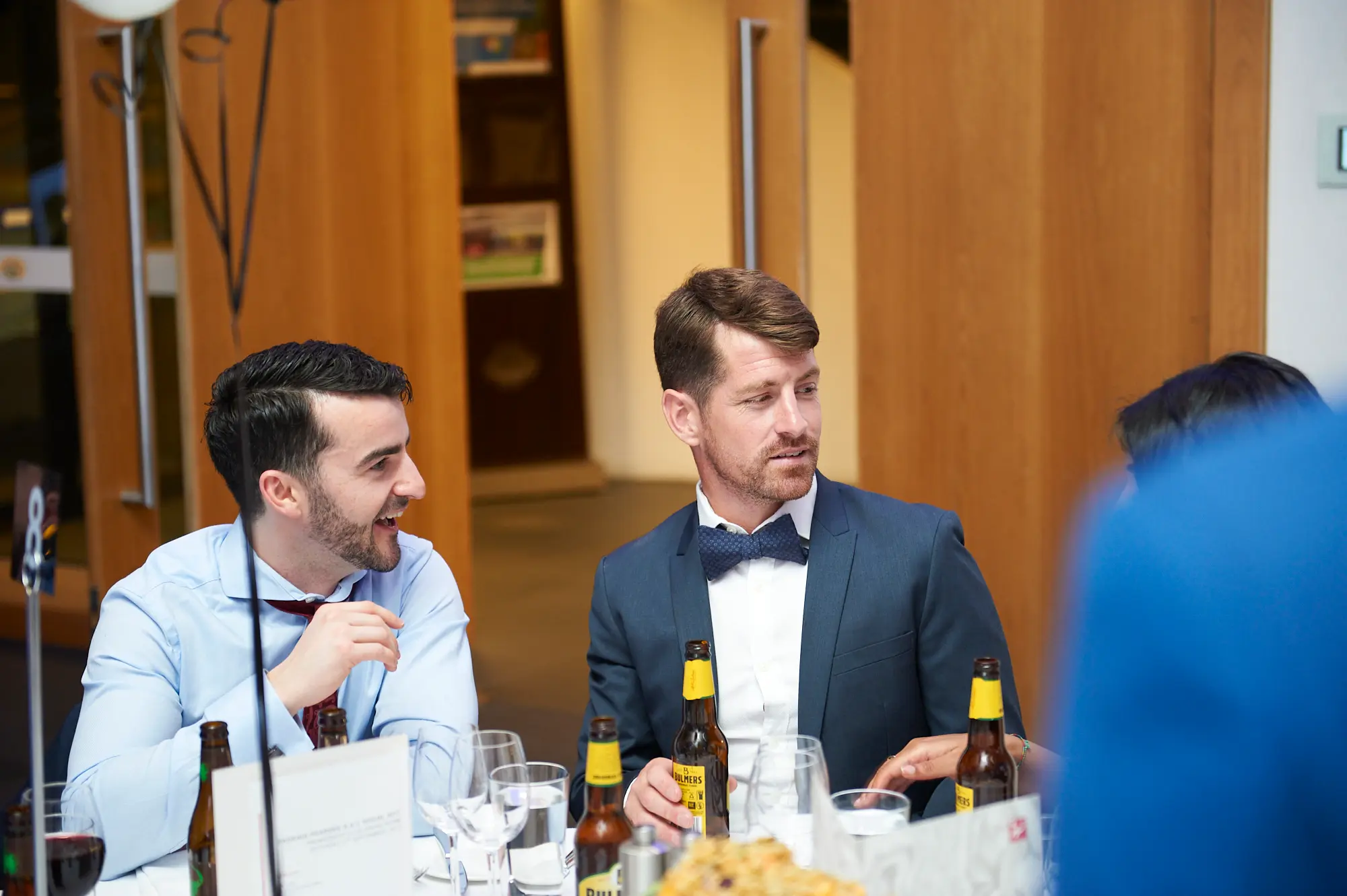 padraig pearses gac awards night mcg candid guests conversing The image captures a candid moment of socializing at the Pearses Awards Night & Gala Dinner at the MCG. Two men seated at a table engage in lively conversation, with the man in a light blue shirt and maroon tie laughing openly, while the man in a dark blue suit and bow tie listens attentively with a relaxed posture. Wine glasses, cutlery, and beer bottles are arranged on the white-clothed table. The background shows modern wooden paneling, a staircase, and warm ambient lighting, highlighting the contemporary venue. The scene conveys a festive, relaxed, and sociable atmosphere during the gala.