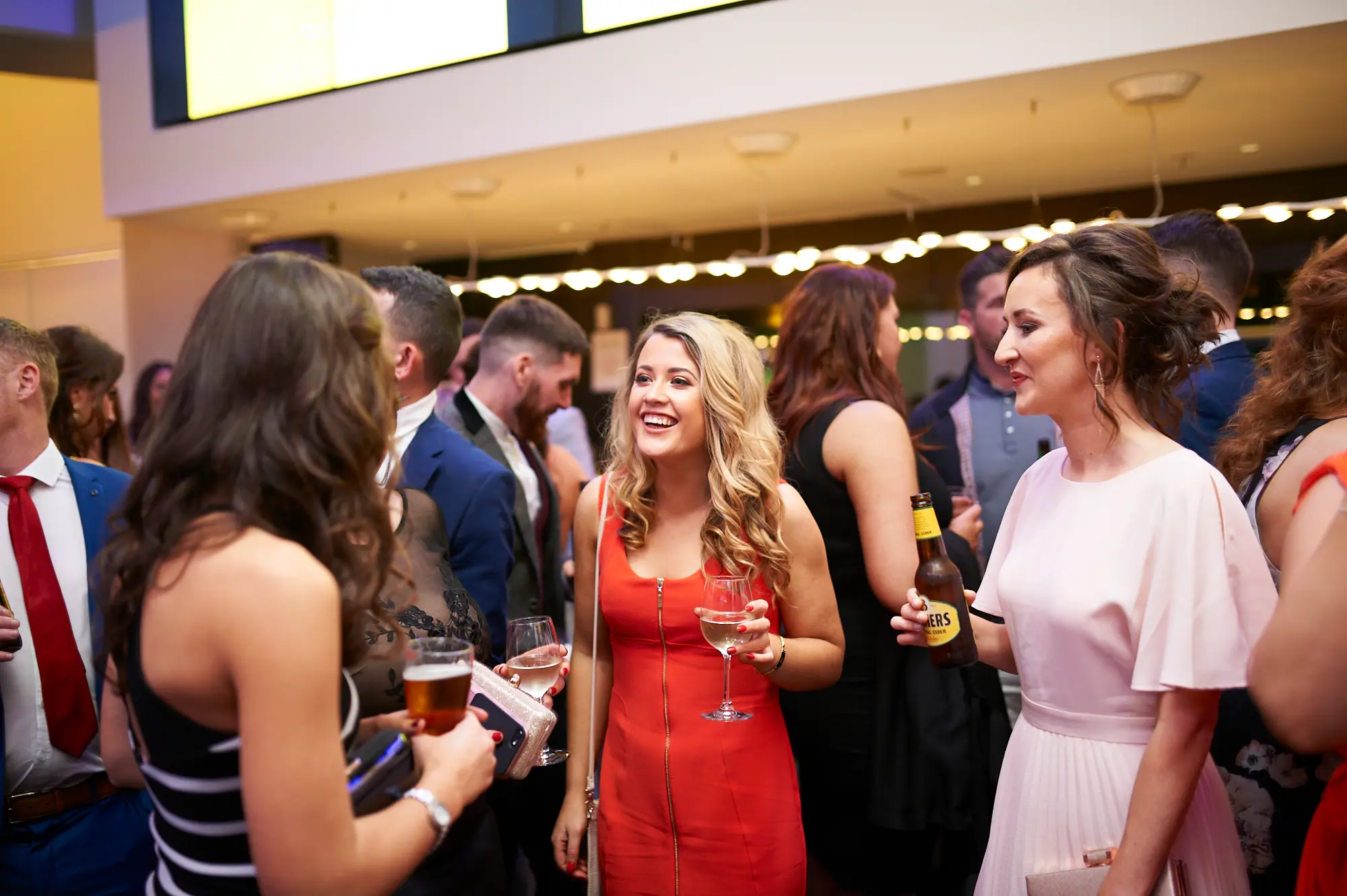 padraig pearses gac awards night mcg candid guests laughing The image captures a candid moment of socializing during the Pearses Awards Night & Gala Dinner at the MCG. A woman in a vibrant red dress laughs heartily while holding a wine glass, joined by a woman in a light pink dress holding a beer, and another guest in a black-and-white striped top, all engaged in lively conversation. Guests in suits and gowns mingle in the blurred background near a bar area. Warm, ambient lighting highlights the foreground, while the contemporary venue with sleek architecture provides a sophisticated backdrop for this spontaneous, festive, and joyful moment.