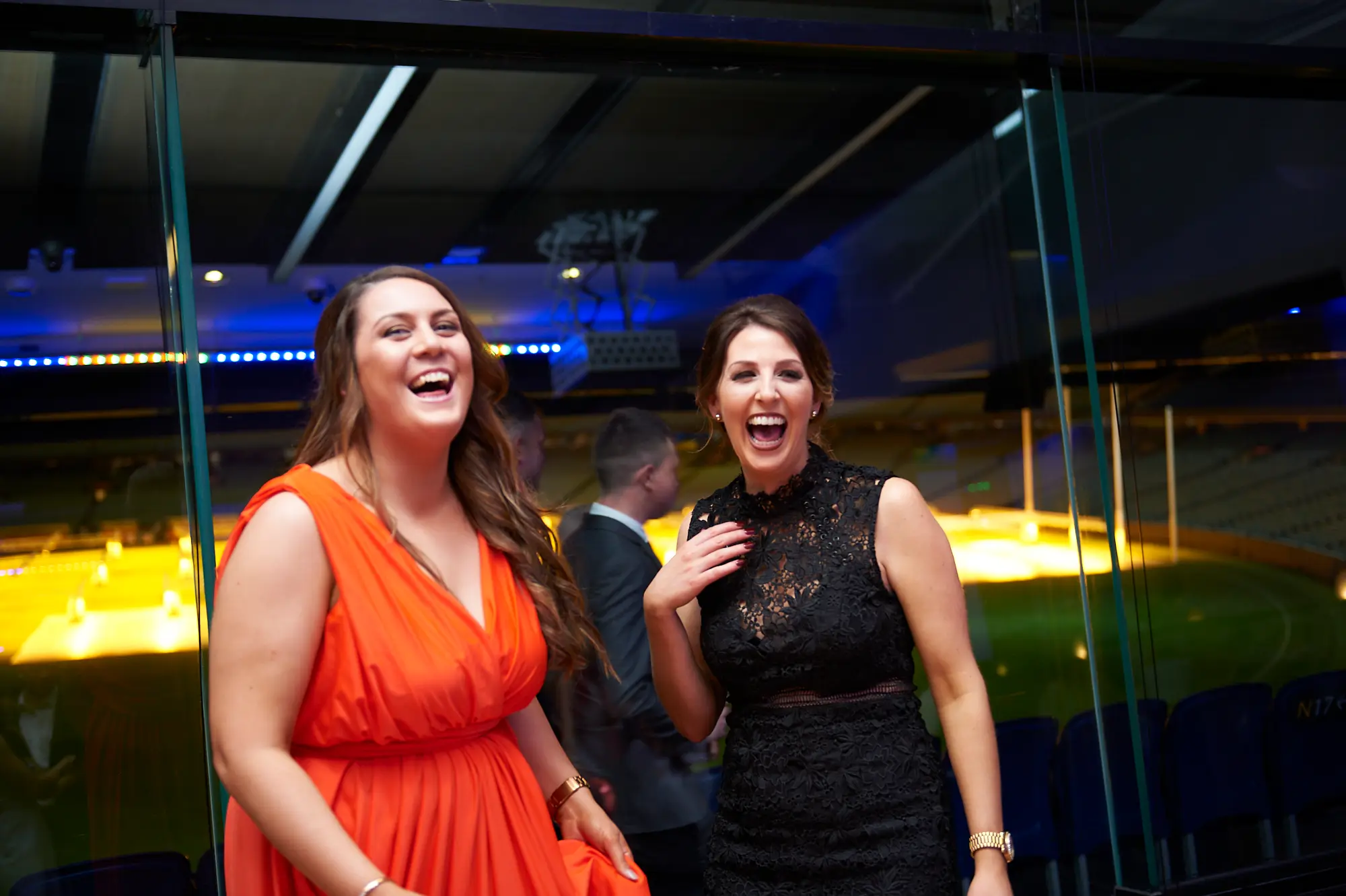 padraig pearses gac awards night mcg candid laughter friends The image captures a candid moment of joyous laughter between two women at the Pearses Awards Night & Gala Dinner at the MCG. One wears a vibrant orange dress, tilting her head back in heartful amusement, while the other in a black lace dress laughs openly, hand near her chest. Behind them, blurred guests mingle, adding to the lively social atmosphere. Large floor-to-ceiling glass partitions reveal dark blue stadium seating and the green MCG field, with warm lighting and vibrant blue ambient reflections. Their genuine expressions convey a festive, celebratory, and energetic mood at the formal gala.