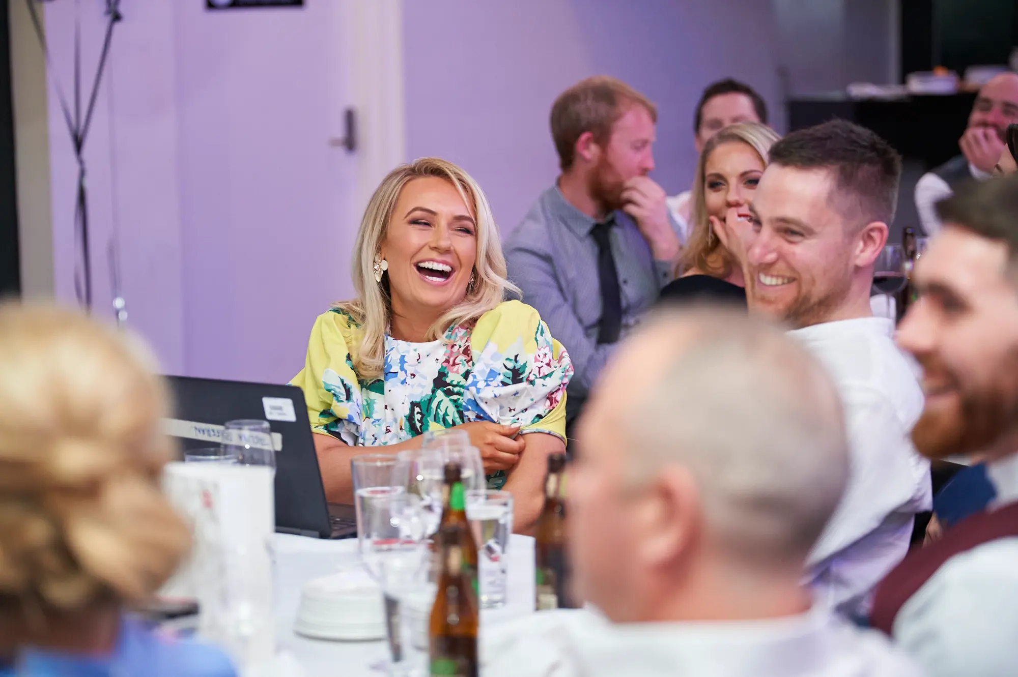 padraig pearses gac awards night mcg candid laughter table The image captures a candid moment of joy and laughter during the Pearses Awards Night & Gala Dinner at the MCG. A blonde woman in a yellow floral dress laughs heartily, head slightly tilted back, while a man in a white shirt smiles broadly toward her. Another woman beside them reacts pleasantly, hand near her mouth. The table is set with wine glasses, cutlery, beer bottles, and a laptop. In the softly lit background, other guests in formal attire are seated or standing, contributing to a lively, social, and celebratory atmosphere at the gala event.
