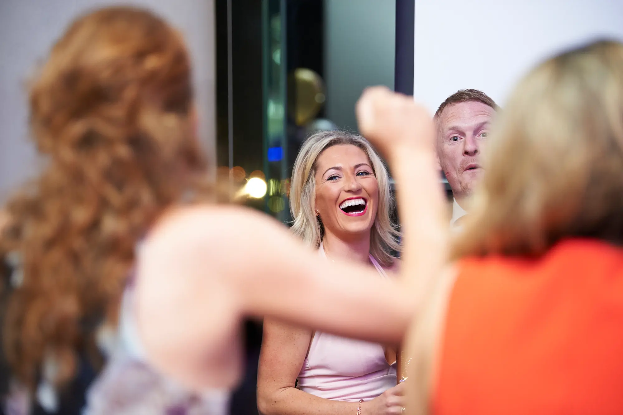 padraig pearses gac awards night mcg candid lively laughter The image captures a candid moment of lively celebration during the Pearses Awards Night & Gala Dinner at the MCG. A woman in a light pink dress laughs heartily with her mouth wide open and bright expression, while a man behind her reacts with wide eyes and an animated face. Blurred figures in the foreground and raised arms add to the sense of energetic social interaction. The dimly lit modern indoor venue, with subtle background lights and dark architectural elements, emphasizes the festive, joyful, and highly social atmosphere of this candid, memorable moment at the gala event.