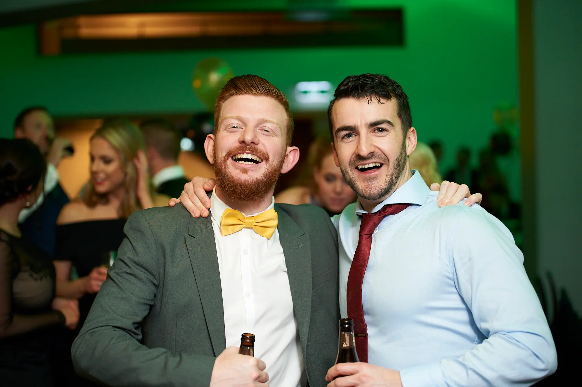 padraig pearses gac awards night mcg candid male friends Two men pose together at the Pearses Awards Night & Gala Dinner at the MCG, arms around each other’s shoulders, laughing openly while holding brown glass beer bottles. They are dressed in a grey suit with a yellow bow tie and a light blue shirt with a red tie. The dark indoor venue features green ambient lighting, gold balloons, and other elegantly dressed guests mingling, capturing a lively, festive, and social gala atmosphere.