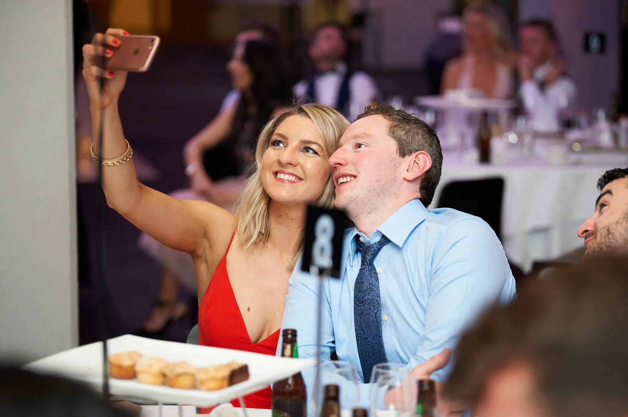 padraig pearses gac awards night mcg candid selfie table The image captures a candid moment of guests taking a selfie at the Pearses Awards Night & Gala Dinner at the MCG. A woman in a vibrant red dress holds her smartphone, smiling broadly at the screen, while a man in a light blue shirt and dark tie leans in, sharing the moment with an engaged smile. A table number "8" sign and a plate of appetizers are visible in the foreground. The background shows other guests seated at round tables with wine glasses and plates, creating a festive, lively, and social atmosphere in the warmly lit event hall.