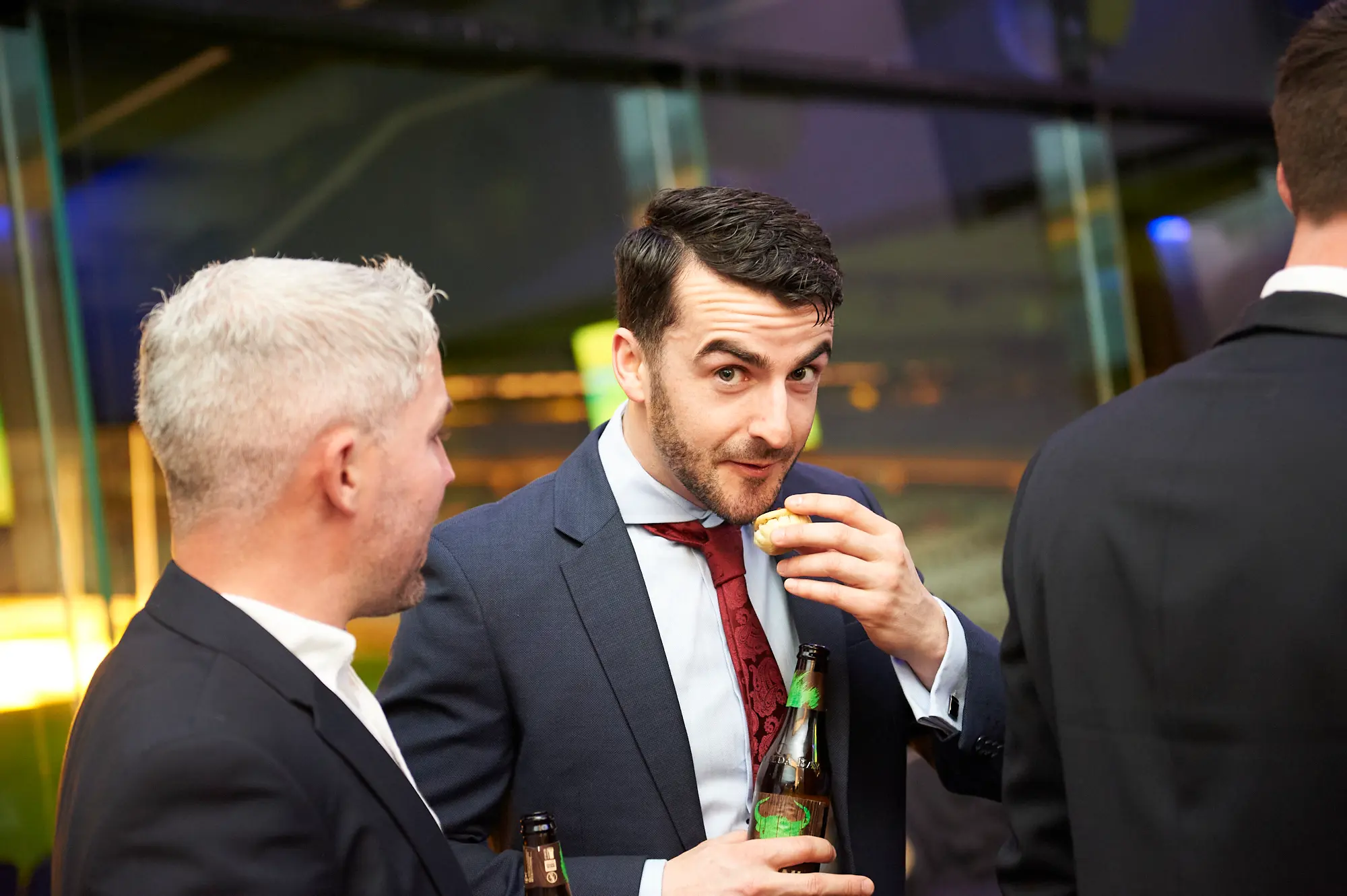 padraig pearses gac awards night mcg candid snacking moment The image captures a candid moment of socializing and casual snacking during the Pearses Awards Night & Gala Dinner at the MCG. A man with a dark beard, dressed in a dark blue suit and red tie, is mid-bite while holding a beer bottle, giving an amused, caught-in-the-moment expression toward the camera. Beside him, a grey-haired man in a black suit jacket looks on with a neutral, attentive expression. Another guest stands with his back to the camera. Behind them, large glass partitions reveal vibrant blue and warm yellow lighting, blurred figures, and a modern, lively event atmosphere.
