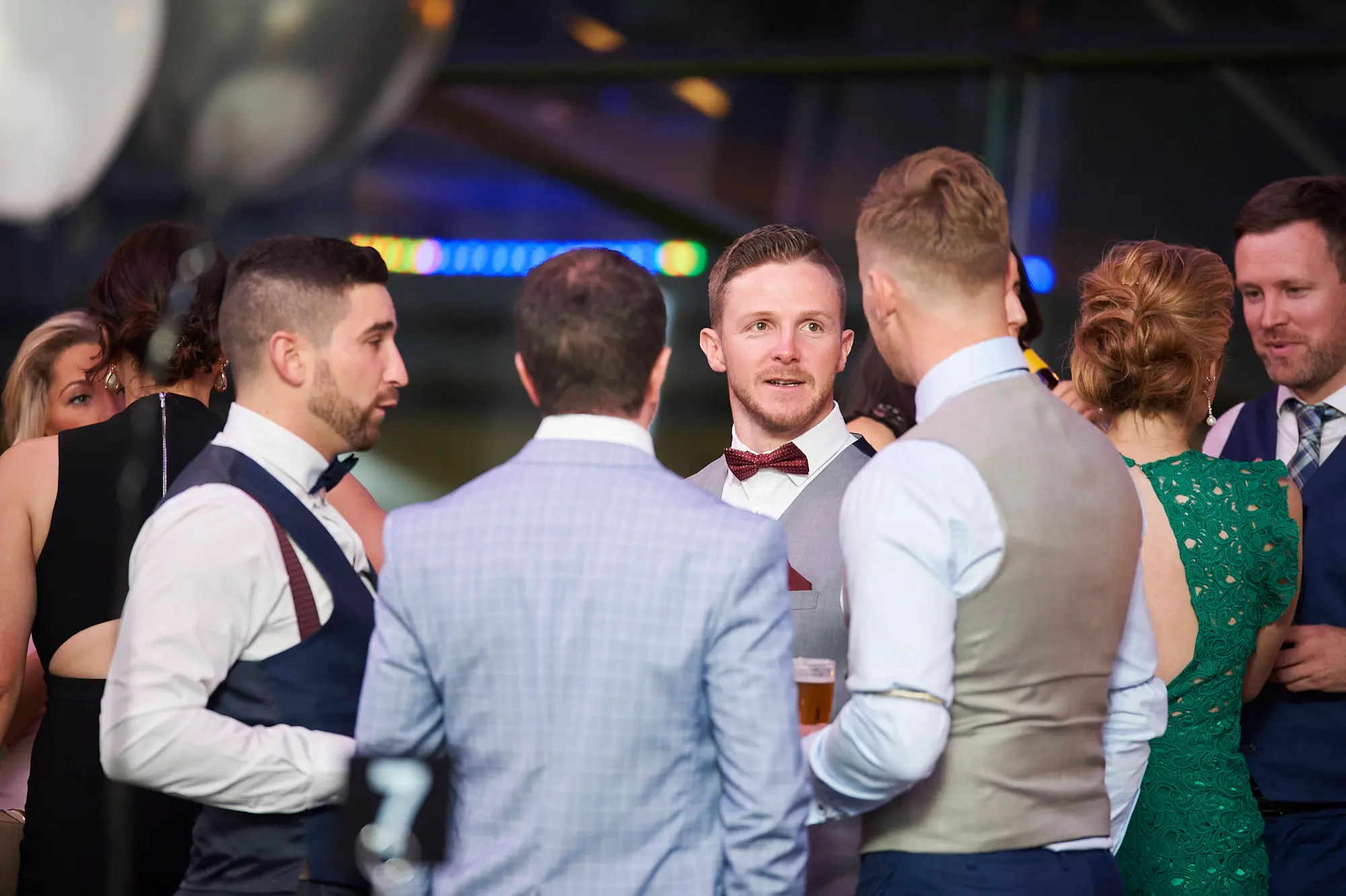 padraig pearses gac awards night mcg candid socializing group The image captures a candid moment of conversation and socializing during the Pearses Awards Night & Gala Dinner at the MCG. A group of men in formal suits and waistcoats, along with two women in evening dresses, are engaged in attentive and animated discussion. One man with a red bow tie looks toward another with a thoughtful expression, while others hold drinks and interact naturally. Dim ambient lighting, blue accent lights, blurred reflections, and floating balloons in the modern stadium function room create a sophisticated yet festive backdrop, emphasizing the relaxed, social, and lively atmosphere of the gala event.