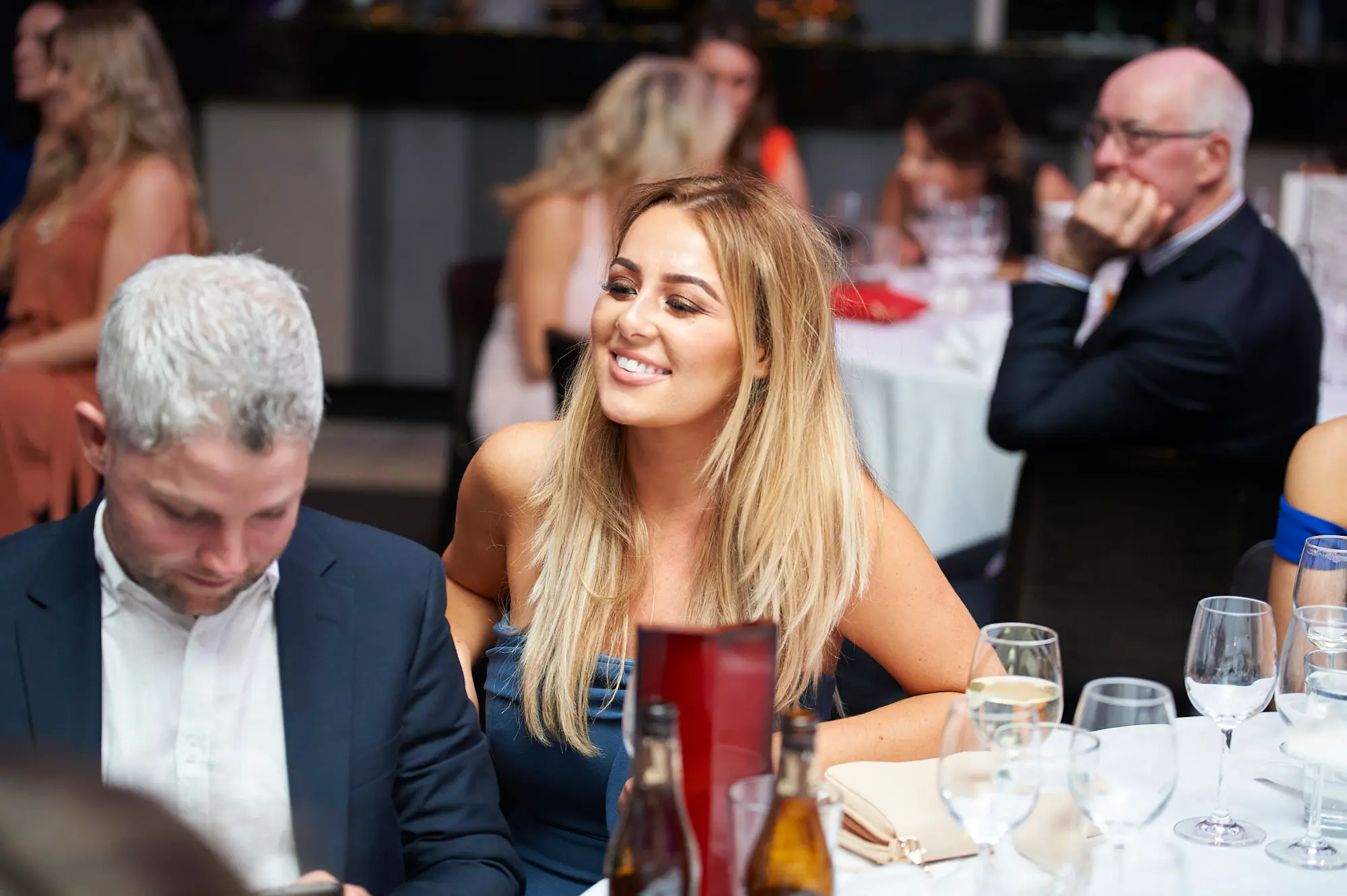 padraig pearses gac awards night mcg candid table smile women The image captures a candid moment of socializing at the Pearses Awards Night & Gala Dinner at the MCG. A blonde woman in a dark blue strapless dress smiles warmly, engaged in conversation, while a man in a navy suit looks down, focused on the table or program. An older man in glasses observes thoughtfully in the background. The table is set with wine glasses, beer bottles, a can of cider, and a red menu card. Other guests in formal attire are seated at round tables throughout the softly lit hall, creating a relaxed, lively, and social atmosphere.