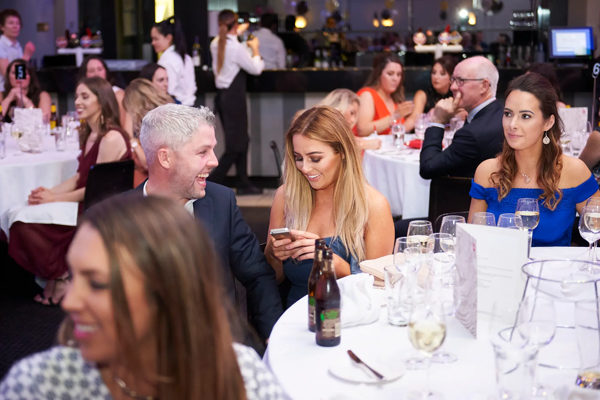 padraig pearses gac awards night mcg candid table socializing The image captures a candid moment of social interaction among guests at the Pearses Awards Night & Gala Dinner at the MCG. A blonde woman in a dark blue dress and a grey-haired man in a navy suit are seated together, smiling and focused on a smartphone, sharing a moment of engagement. Another woman in a vibrant blue off-the-shoulder dress looks to the side with a pleasant expression, wine glass in front of her. In the background, elegantly dressed guests sit at round tables, and staff attend a bar, creating a lively, relaxed, and festive gala atmosphere.