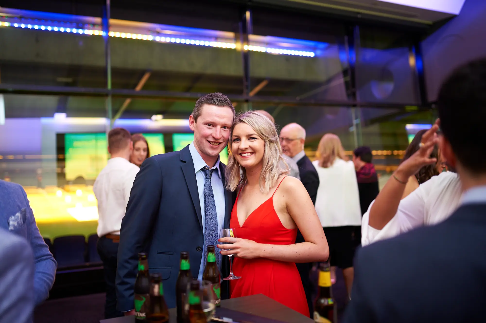 padraig pearses gac awards night mcg couple arrival red dress A couple poses warmly during the arrival of the Padraig Pearses GAC Awards Night & Gala Dinner at the MCG. The man wears a dark blue suit and tie, holding a small beer bottle, while the woman wears a vibrant red spaghetti-strap dress and holds a wine glass. Their close, happy pose conveys shared enjoyment. The modern event space features floor-to-ceiling glass windows, blue and green ambient lighting, and a bar counter with drinks. Other formally dressed guests mingle in the blurred background, creating a vibrant, elegant, and welcoming atmosphere as the evening’s gala festivities begin.