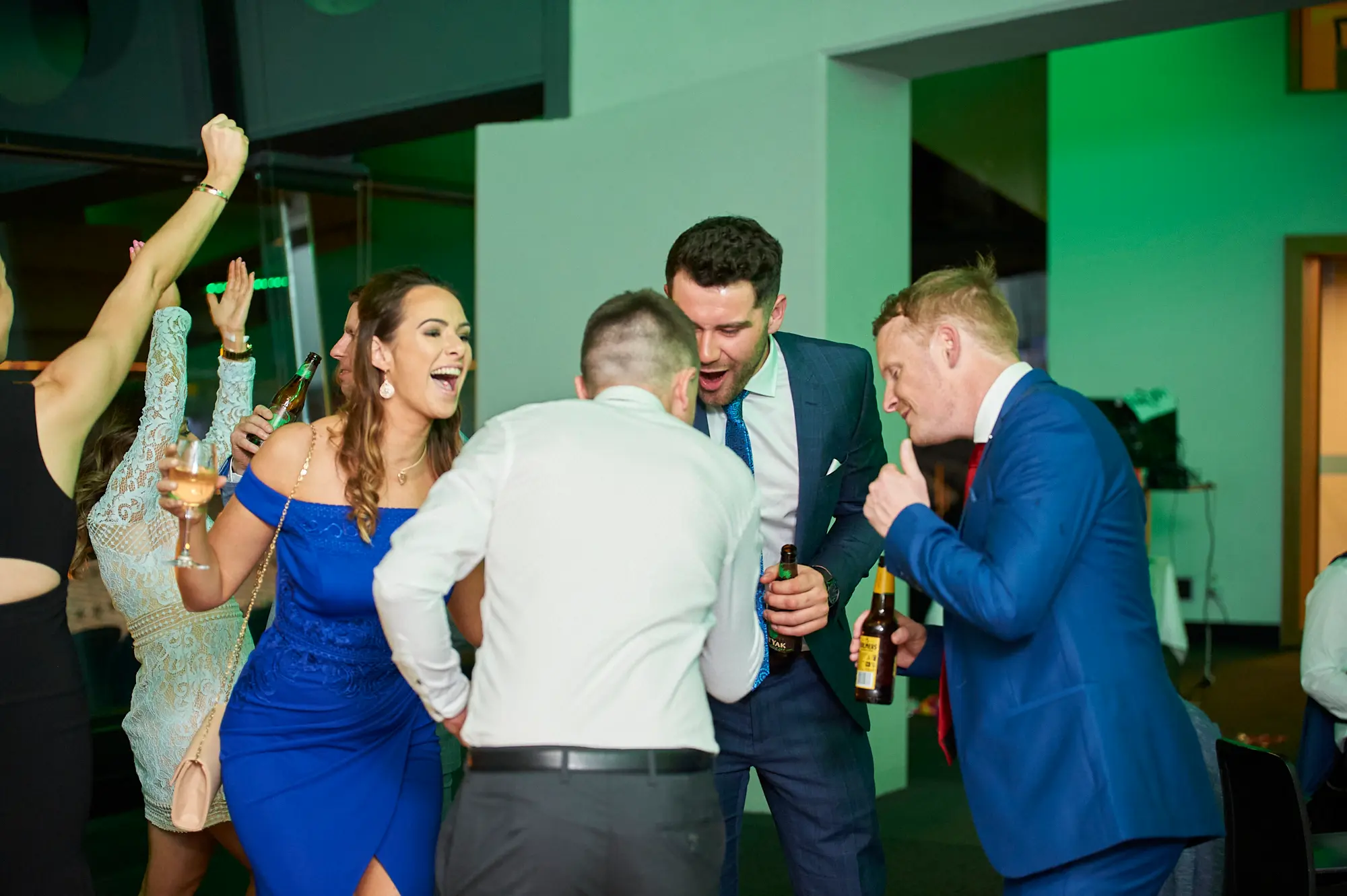 padraig pearses gac awards night mcg dance floor celebration fun Guests celebrate energetically on the dance floor at the Pearses Awards Night & Gala Dinner at the MCG. A bearded man in a white shirt laughs ecstatically with a beer bottle, while a man in a blue suit points enthusiastically at him. Two women, one in a vibrant blue off-the-shoulder dress and another in a white lace dress, cheer and laugh, holding a clutch and wine glass. Surrounded by vibrant green ambient lighting and glass partitions revealing stadium seating, the group’s animated expressions capture the lively, joyful, and social atmosphere of the gala’s party phase.