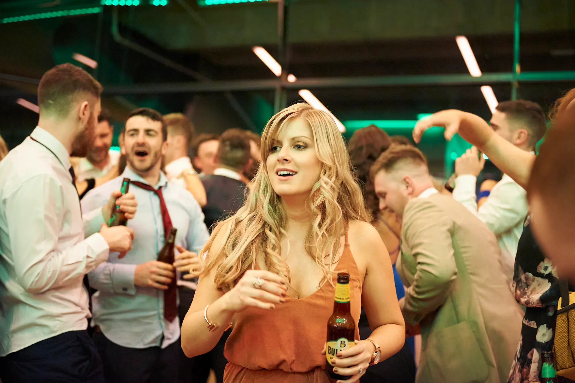 padraig pearses gac awards night mcg dance floor celebration Guests celebrate energetically on the dance floor at the Pearses Awards Night & Gala Dinner at the MCG. A woman in an orange dress smiles holding a beer, while a man beside her laughs openly, surrounded by dancing attendees in formal attire. Vibrant green lighting and a modern venue backdrop create a lively, festive atmosphere filled with joy, camaraderie, and the spirited energy of the night.