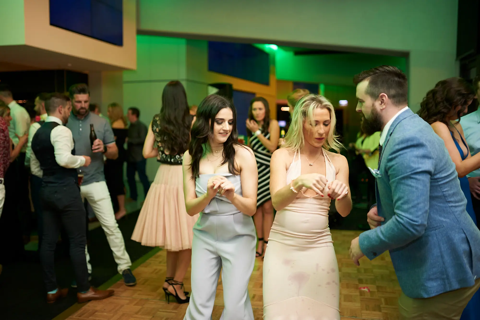 padraig pearses gac awards night mcg dance floor energy two women Guests celebrate energetically on the dance floor during the Pearses Awards Night at the MCG, with two women in light blue and pink/champagne dresses laughing and gesturing mid-dance. A man in a blue suit leans in, smiling, while other guests mingle and clap nearby. The modern function room glows with vibrant green ambient lighting, reflecting off sleek architectural elements and floor-to-ceiling glass partitions. Wooden dance flooring anchors the scene, and the overall atmosphere captures the excitement, camaraderie, and lively energy of a formal gala that has transformed into a joyful, interactive party.