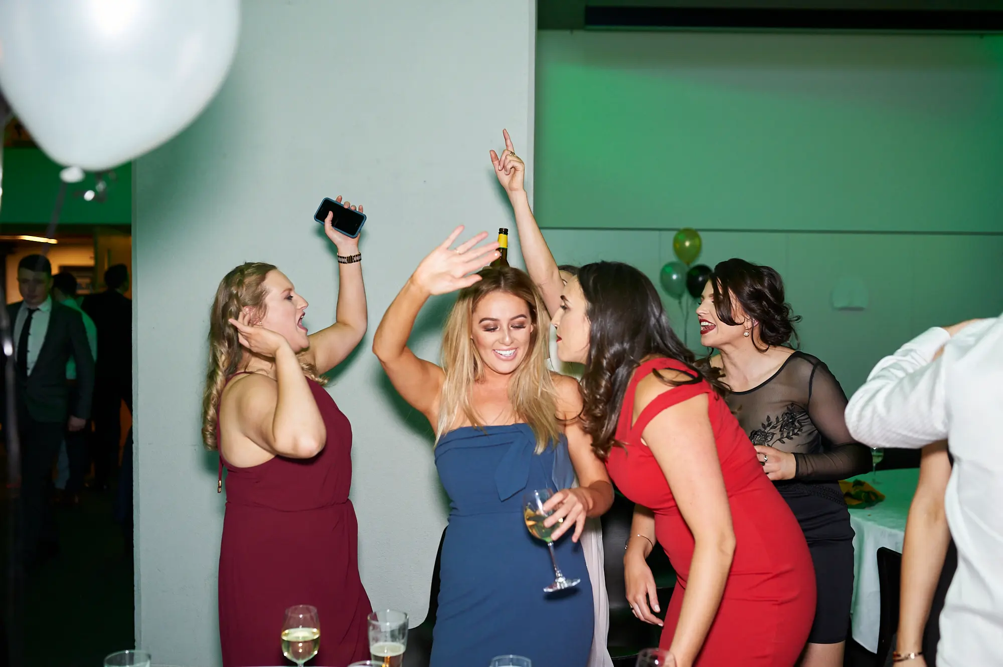 padraig pearses gac awards night mcg dance floor energy Four women on the dance floor at the Pearses Awards Night & Gala Dinner at the MCG laugh, cheer, and raise their arms in excitement. The woman in maroon holds a smartphone, the blonde in dark blue holds a wine glass, and the woman in red leans in playfully. Guests in formal attire mingle around under vibrant green lighting, with festive balloons adding to the lively, celebratory atmosphere of the gala.
