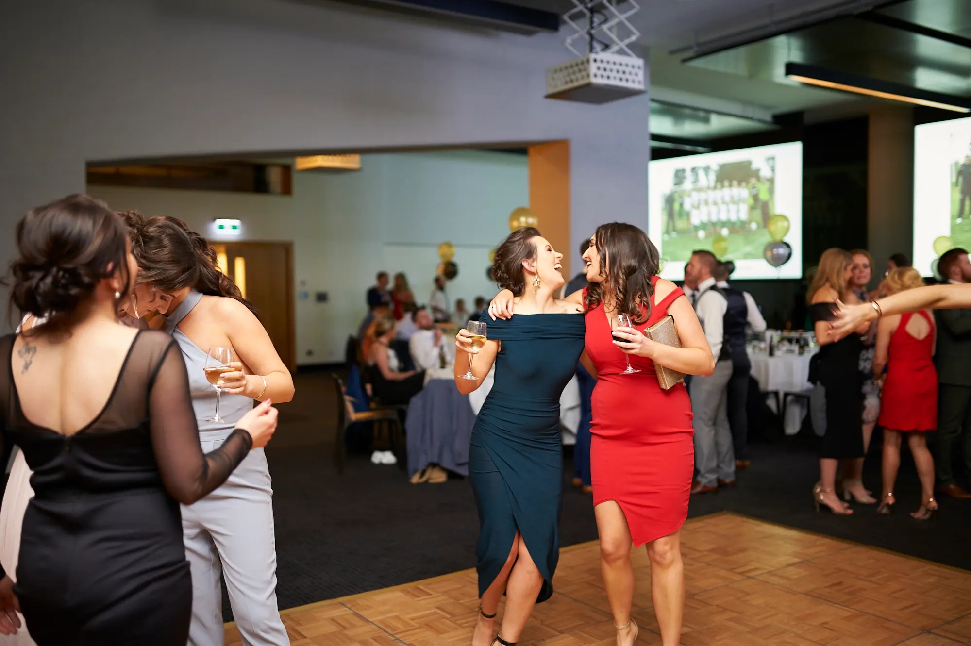 padraig pearses gac awards night mcg dance floor fun Two women in elegant evening gowns laugh and interact on the dance floor at the Pearses Awards Night & Gala Dinner at the MCG. The woman in red smiles broadly while holding a wine glass, and the woman in teal laughs heartily, capturing spontaneous joy. Guests in formal attire mingle around, with ambient green and blue lighting and a wooden dance floor enhancing the lively, celebratory atmosphere of the night.