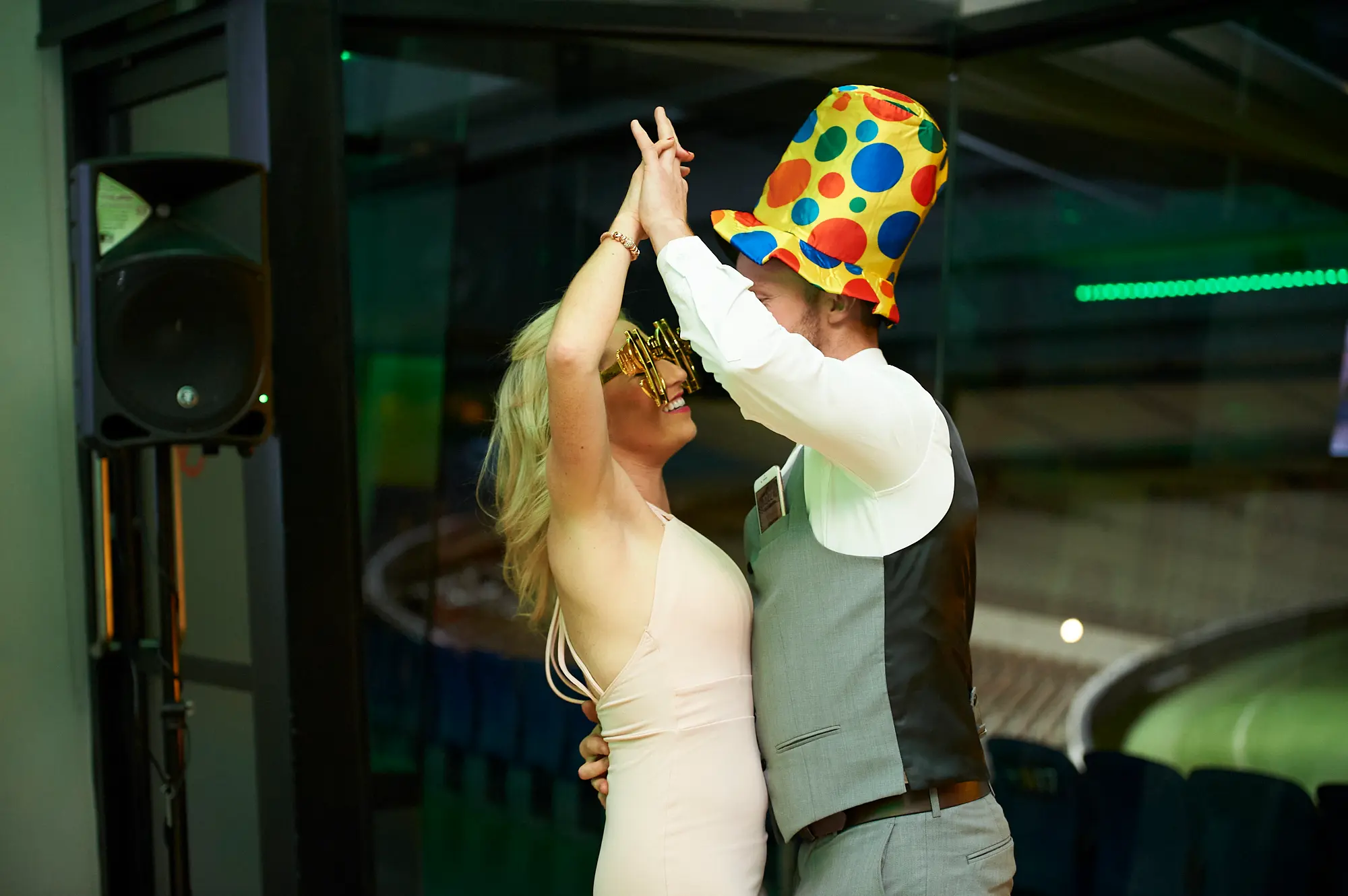 padraig pearses gac awards night mcg dance floor spin A man in a grey waistcoat and white shirt wearing a colorful polka-dot party hat dances energetically with a woman in a light slip dress and oversized gold-framed glasses at the Pearses Awards Night & Gala Dinner at the MCG. Holding hands high, they smile at each other mid-spin on the dance floor, surrounded by vibrant green and blue lighting, with guests and stadium seating visible through glass partitions in the background, capturing the lively, playful, and celebratory atmosphere of the gala’s party phase.
