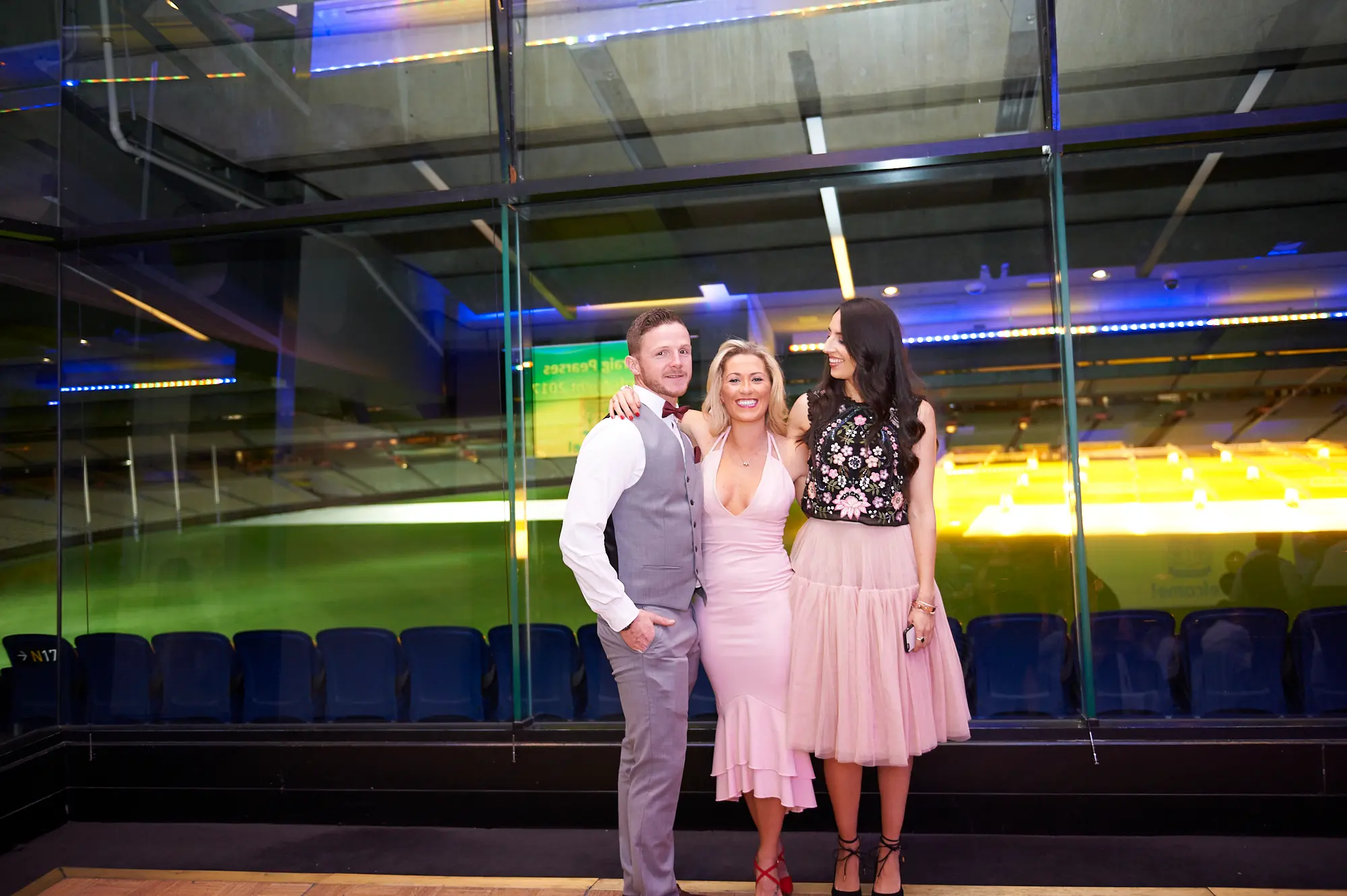 padraig pearses gac awards night mcg elegant group posing Three guests pose together at the Pearses Awards Night & Gala Dinner at the MCG, smiling warmly at the camera. A man in a grey waistcoat stands with arms around two women in light pink and black/floral dresses. They are elegantly dressed and relaxed, with floor-to-ceiling glass partitions behind them revealing dark blue stadium seating and the green MCG field, accented by vibrant blue lighting, capturing a sophisticated and festive gala atmosphere.