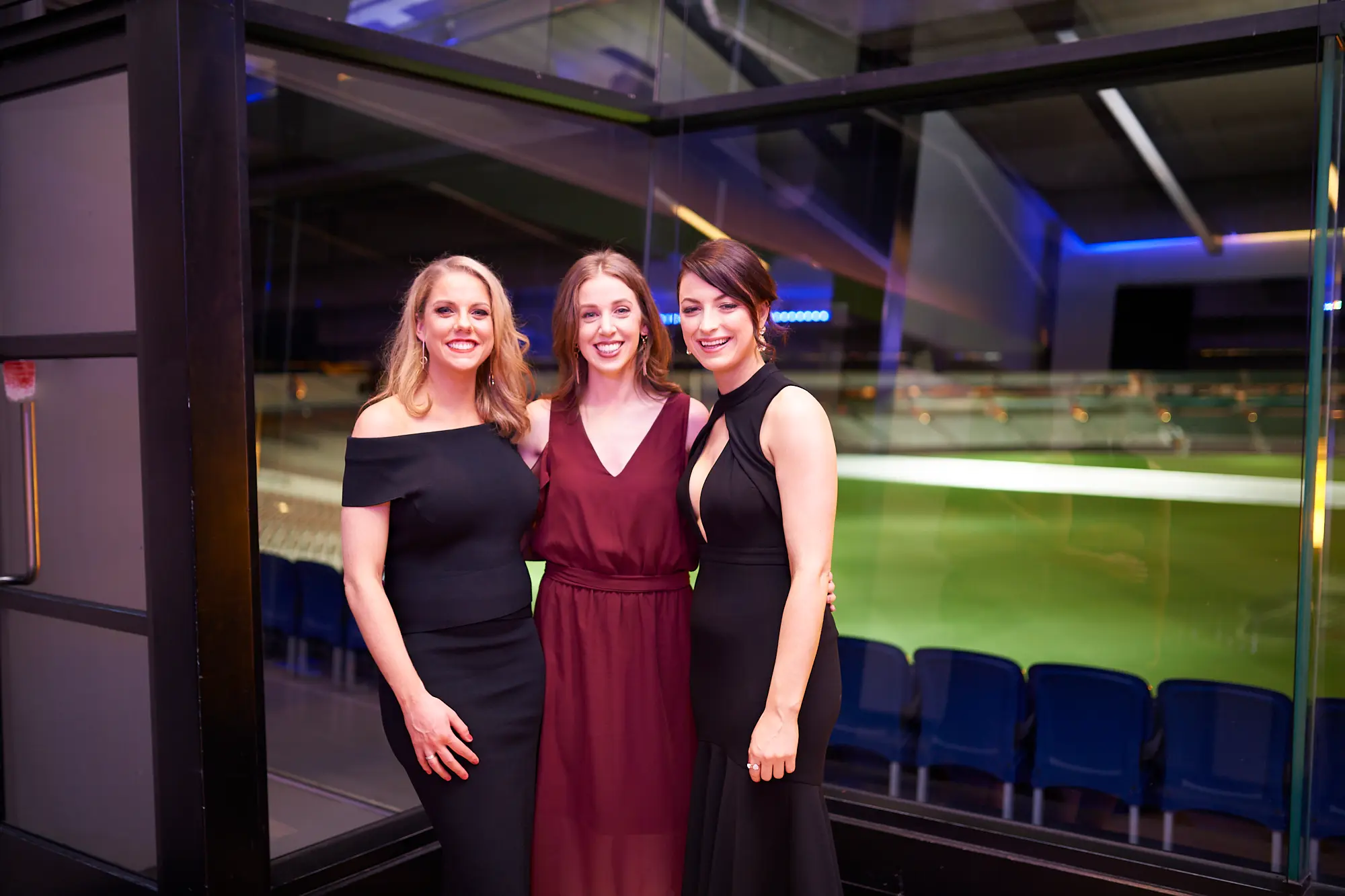 padraig pearses gac awards night mcg elegant posing Three women pose together during the Pearses Awards Night at the MCG, dressed in elegant evening gowns—black off-shoulder, burgundy V-neck, and black high-neck—smiling warmly at the camera. They stand in a modern function room with floor-to-ceiling glass partitions showing the dark blue stadium seating and green MCG field beyond. Vibrant blue accent lighting reflects off the glass, highlighting the sophisticated setting. Their relaxed postures and friendly expressions capture the social, celebratory, and elegant atmosphere of the gala event.