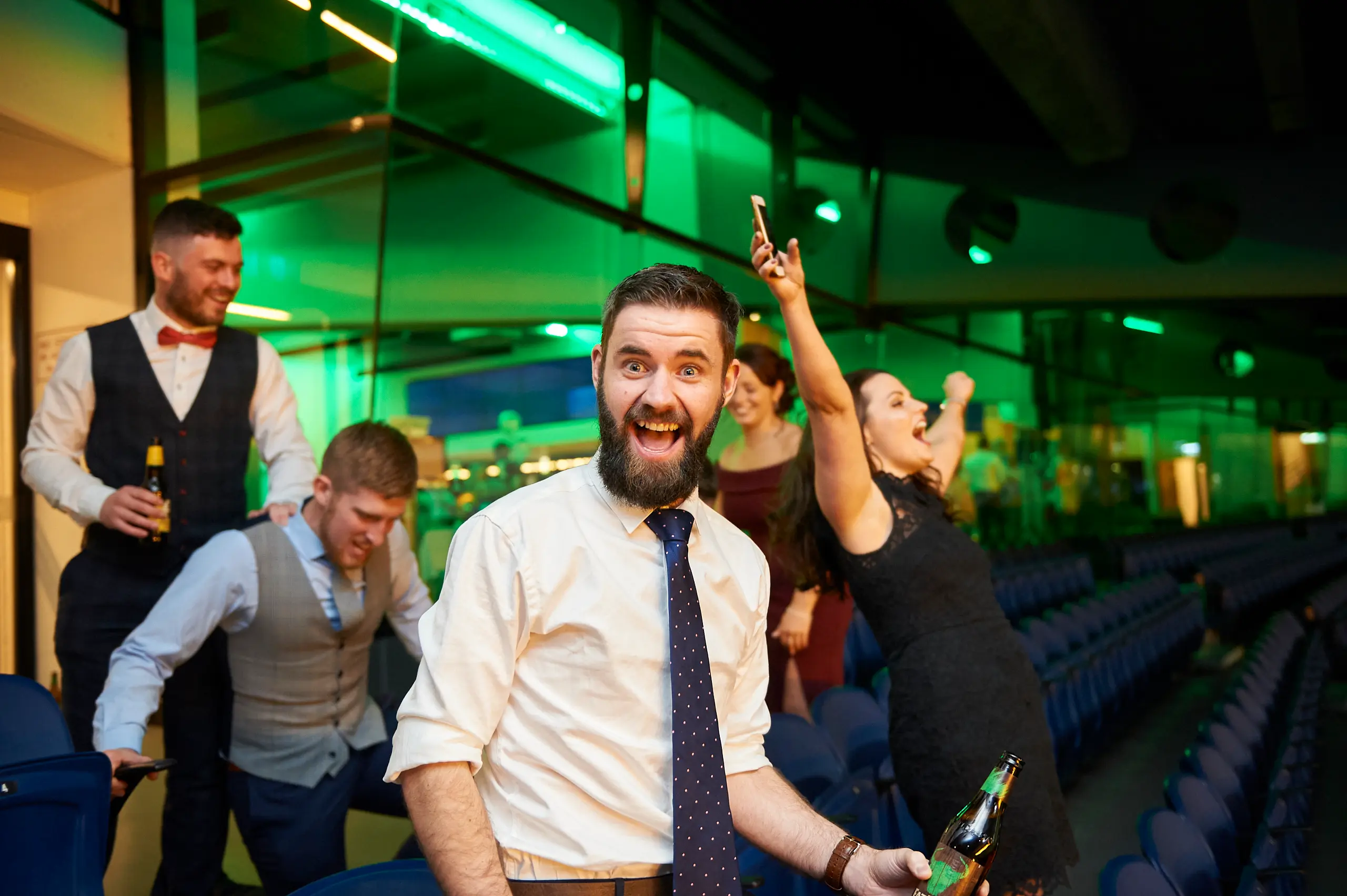 A high-energy celebration erupts at the Padraig Pearses GAC Awards Night & Gala Dinner at the MCG, where guests cheer beneath vibrant green lighting. A bearded man in a white shirt lifts his hand in excitement while holding a beer, as a woman beside him raises her phone to capture the moment. Surrounded by formally dressed guests joining in the lively atmosphere, the scene reflects the joyful, uninhibited spirit of the event’s after-party.