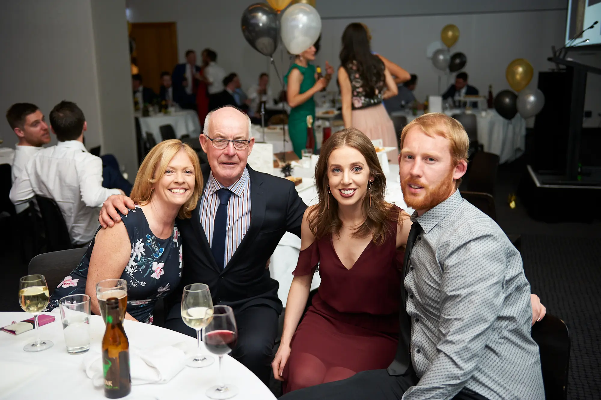 padraig pearses gac awards night mcg family dinner table burgundy A family group shares a joyful moment at a white-clothed dinner table during the Padraig Pearses GAC Awards Night & Gala Dinner at the MCG. An older couple sits on the left, smiling warmly with the man’s arm around the woman, while a younger woman in a burgundy off-the-shoulder dress and a young man in a grey shirt sit on the right, also smiling at the camera. The table features wine glasses, a beer bottle, plates, and cutlery. Festive balloons in black, gold, and white float above tables, while warm ambient lighting highlights the lively, celebratory, and social atmosphere of the gala event.