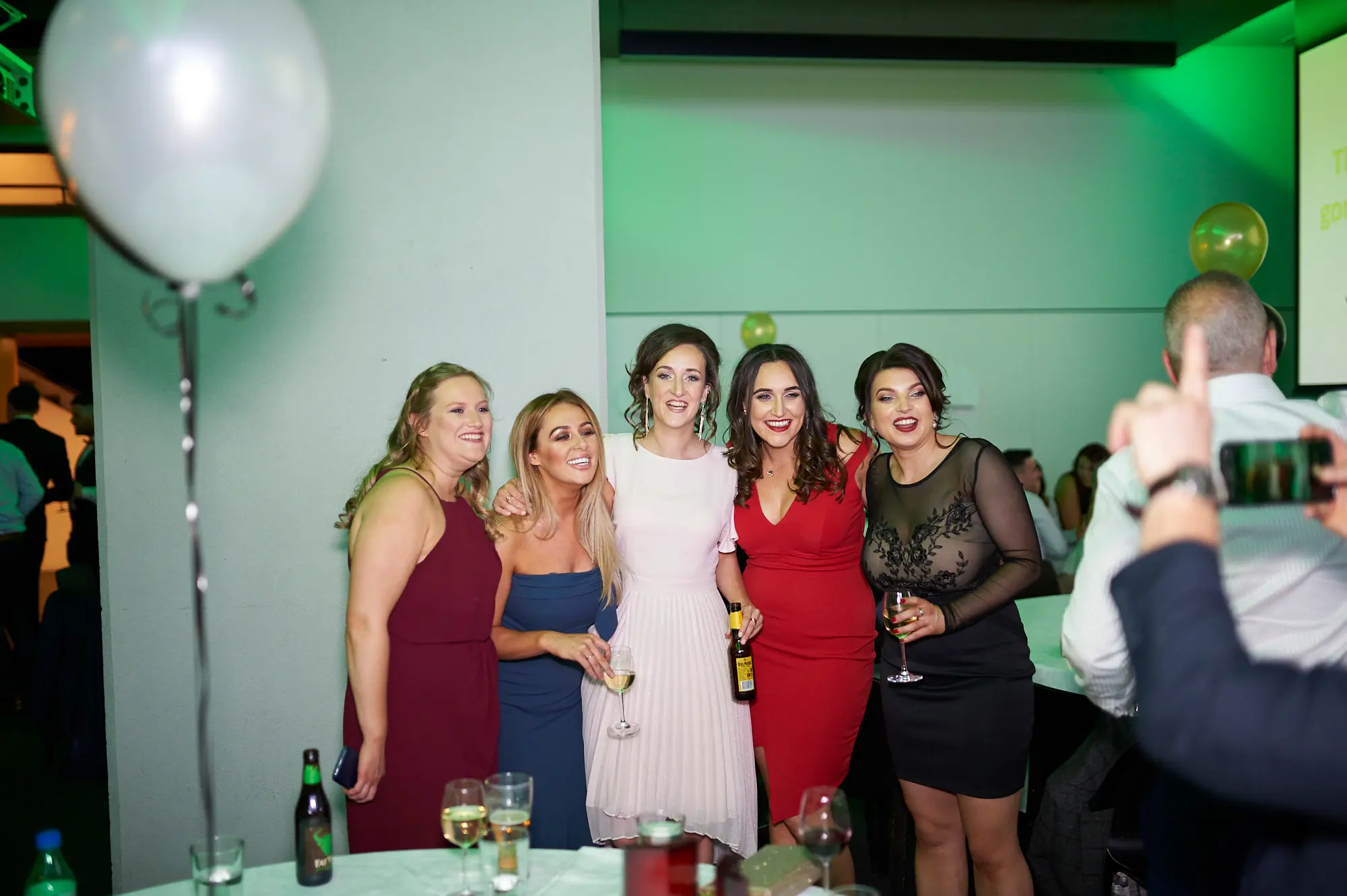 padraig pearses gac awards night mcg female group celebration Five women pose energetically at the Pearses Awards Night & Gala Dinner at the MCG, laughing and smiling while holding wine glasses. Dressed in light pink, dark blue, red, and black lace dresses, they convey camaraderie and joy. Green ambient lighting, gold and white balloons, and other guests in the background create a lively, festive, and social gala atmosphere, while a man captures the moment on his smartphone.