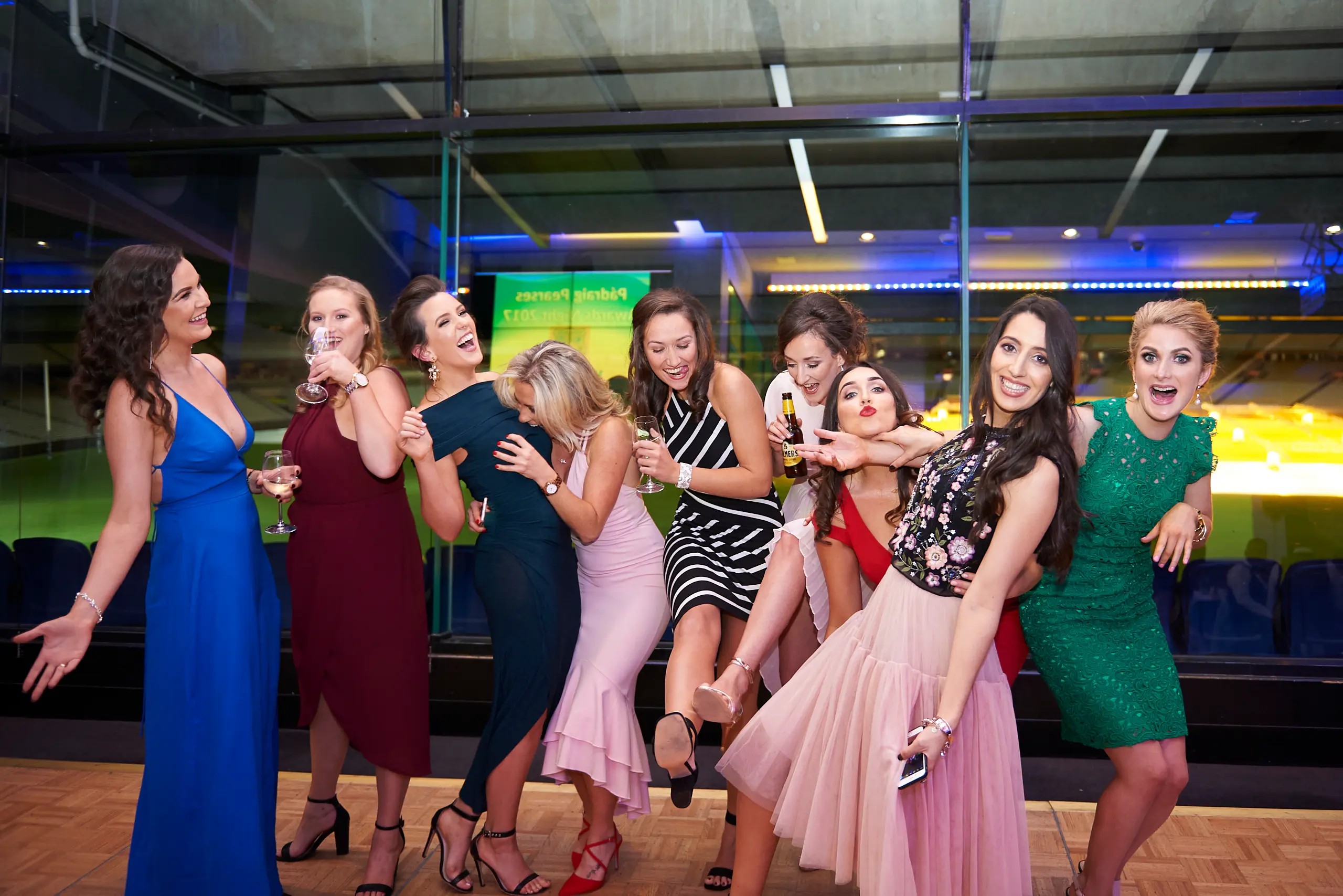 padraig pearses gac awards night mcg friends celebrating laughter A lively group of friends shares a joyful moment at the Padraig Pearses GAC Awards Night & Gala Dinner at the MCG. Women in colorful evening gowns, including dark teal, red, pink, and striped outfits, laugh and toast with drinks, capturing genuine camaraderie. The warm indoor lighting highlights their dynamic expressions against large glass windows revealing stadium seating and the MCG field outside, blending formal elegance with playful celebration and creating a memorable, energetic atmosphere.