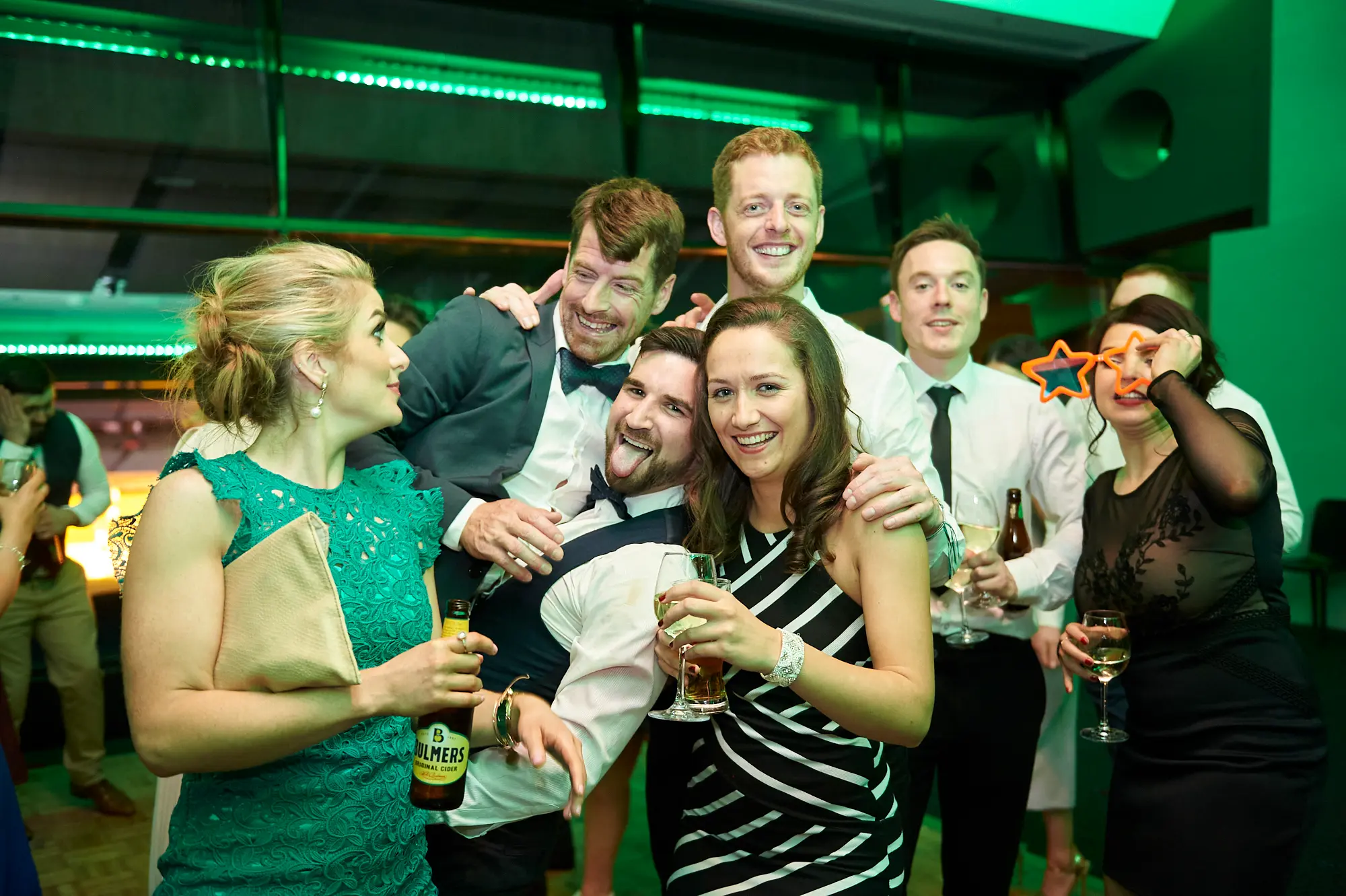 padraig pearses gac awards night mcg fun candid Guests enjoy a lively moment at the Pearses Awards Night at the MCG, with a woman in a green lace dress holding a Bulmers beer and smiling, and a man in a white shirt and waistcoat playfully sticking his tongue out. Nearby, a woman in black lace holds star-shaped glasses over her eyes while holding a wine glass, and other guests hold drinks and smile. The modern function room with floor-to-ceiling glass partitions shows dark stadium seating beyond, while vibrant green ambient lighting adds a dynamic, celebratory atmosphere of shared fun and joyous interaction.