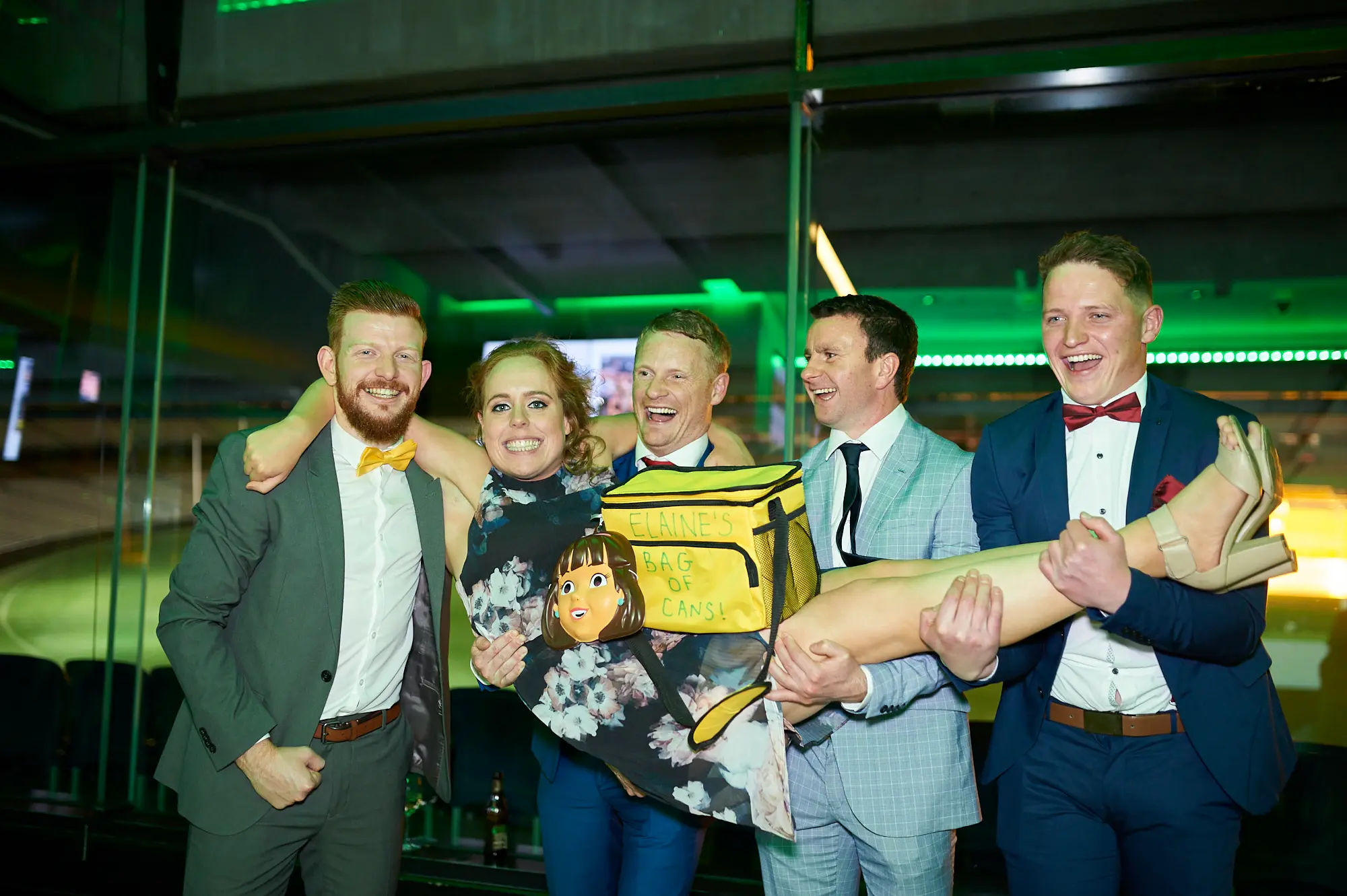 padraig pearses gac awards night mcg fun group with elaine bag Four guests at the Pearses Awards Night & Gala Dinner pose together with laughter and playful energy, holding a custom “ELAINE’S BAG OF CANS.” One man humorously lifts a leg prop while another has an arm around a woman in a floral dress. Dressed in formal attire, they stand in a modern MCG function room with glass partitions, stadium seating visible beyond, and vibrant green ambient lighting reflecting off the walls. The image captures their joyful camaraderie, festive spirit, and shared excitement during a lively, celebratory moment of the gala event.