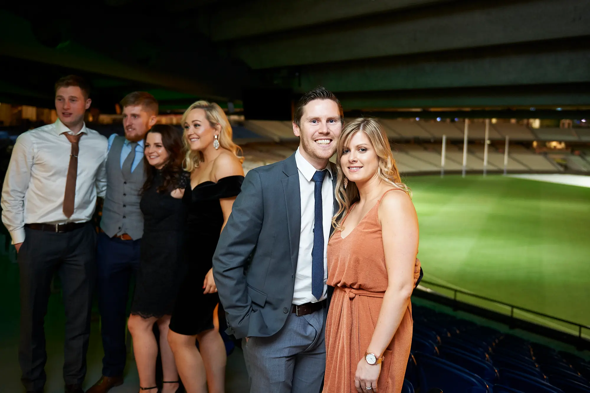 padraig pearses gac awards night mcg group celebration posing Six guests pose together at the Pearses Awards Night at the MCG, dressed in formal attire. In the foreground, a man in a grey suit and a woman in an orange dress smile warmly with arms around each other, while four guests in the background, including women in black dresses, also pose and laugh. The modern venue features large glass partitions revealing dark blue stadium seating and the green MCG field beyond. Vibrant green ambient lighting reflects off the glass, enhancing the festive, elegant, and social atmosphere as the group enjoys the gala event.