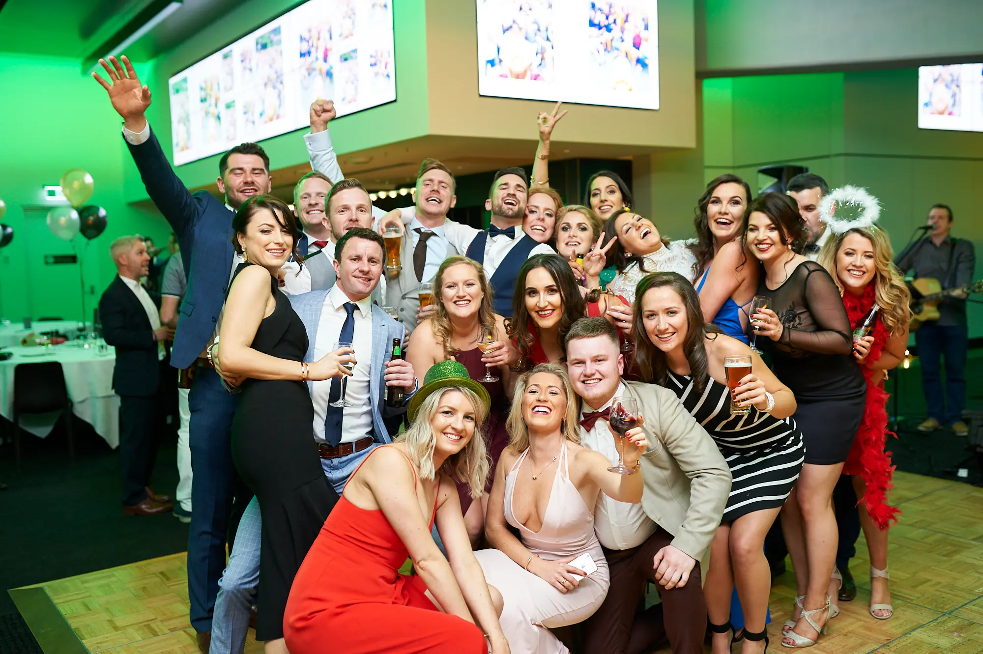 padraig pearses gac awards night mcg group celebration Over 15 gala guests gather closely for a lively group photo during the Pearses Awards Night at the MCG. Men in suits and women in colorful evening gowns smile, laugh, and cheer, some raising hands or holding drinks. The modern function room features a wooden dance floor, vibrant green ambient lighting, large screens, and a bar or stage area with live music. The scene captures energetic camaraderie, playful poses, and a festive, celebratory atmosphere, highlighting the joy and memorable moments shared during the gala event.